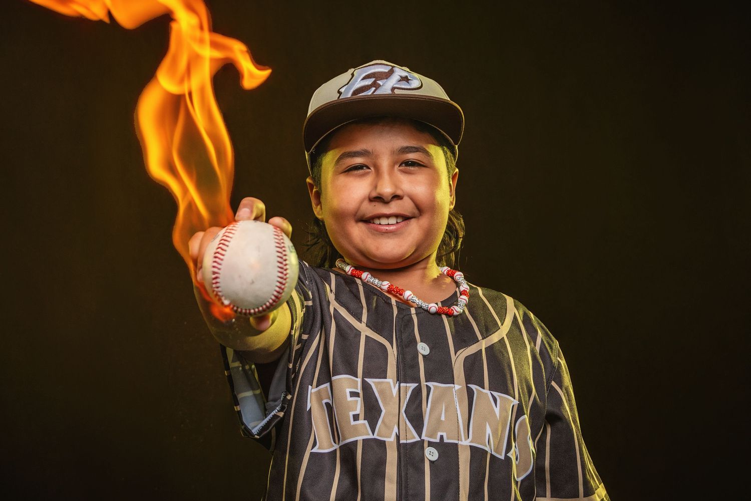 Child in Texans baseball jersey holds a flaming baseball, smiling confidently against a dark background.