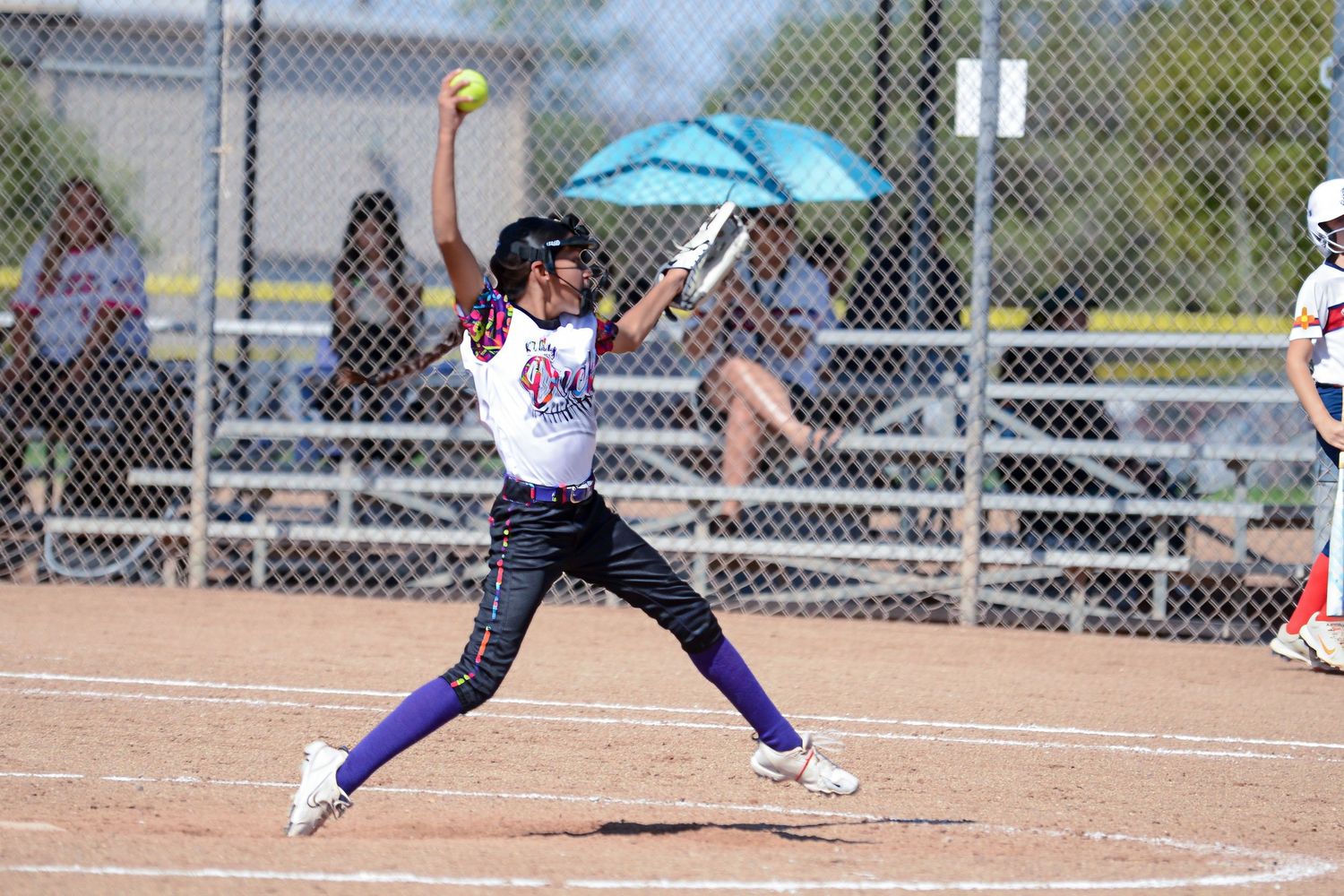 Youth softball pitcher in purple uniform delivers an underhand pitch during a sunny game.