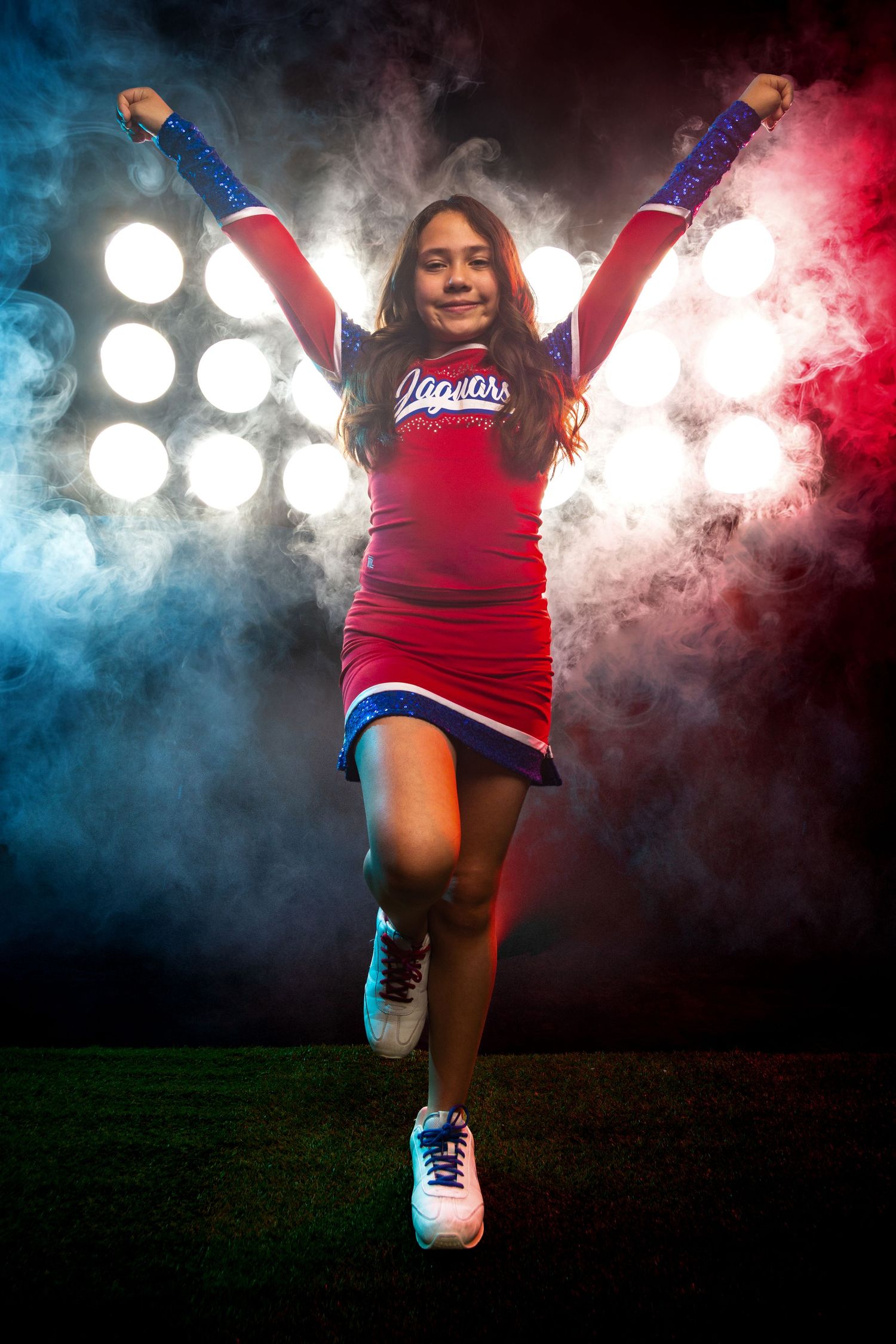 A young cheerleader in a red Jaguars uniform poses with arms raised against dramatic stadium lights and colorful smoke.