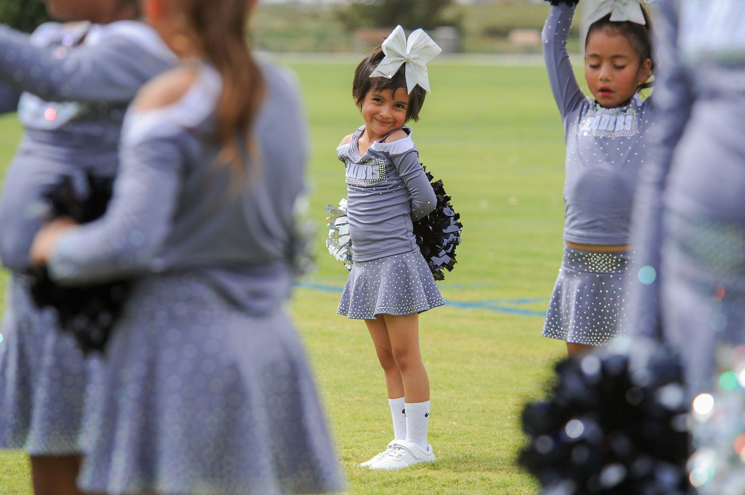 Young girls in gray cheerleader uniforms perform with pom-poms on a green field during a youth cheerleading event.