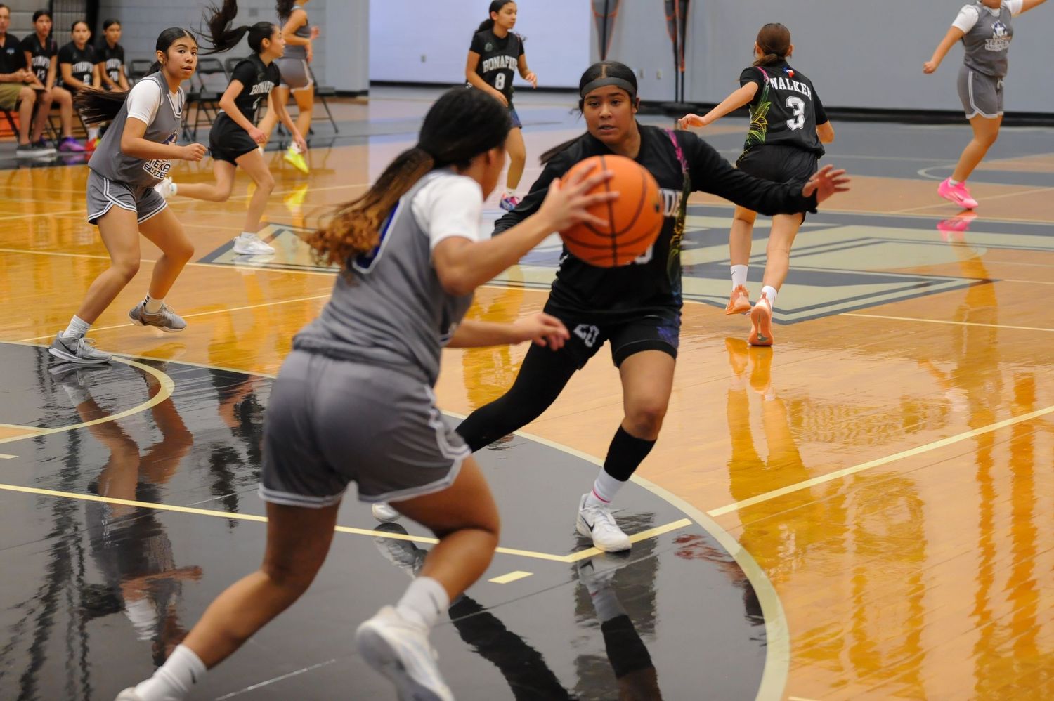 Female high school basketball players practice dribbling and defense drills on an indoor court during a team scrimmage.