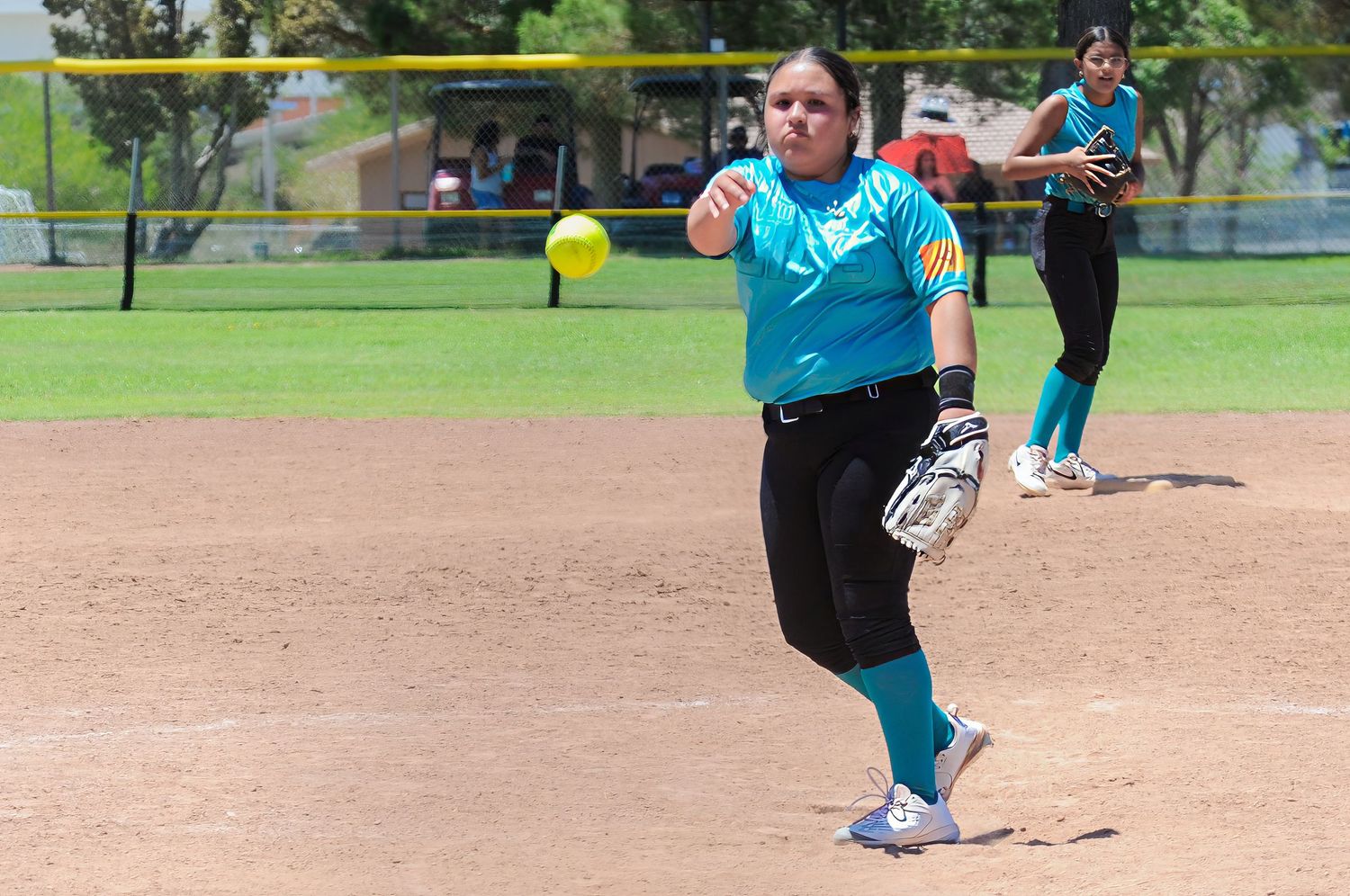 Female softball pitcher in teal uniform releasing a yellow ball during a sunny outdoor softball game.