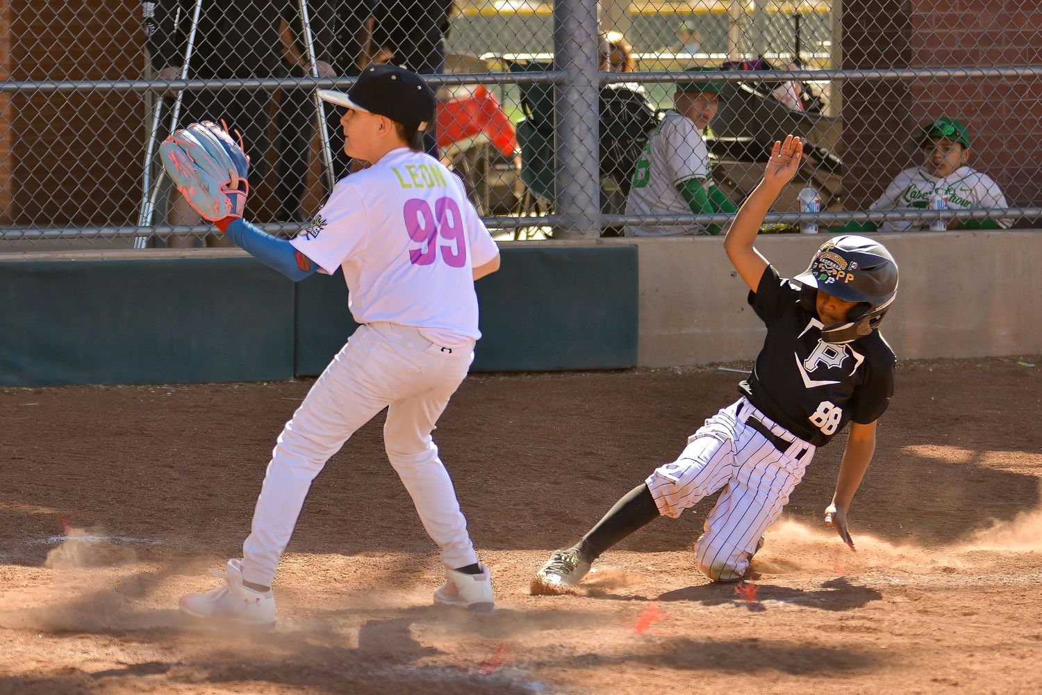 Youth baseball player slides into base as number 99 stretches to catch the ball in a close play.