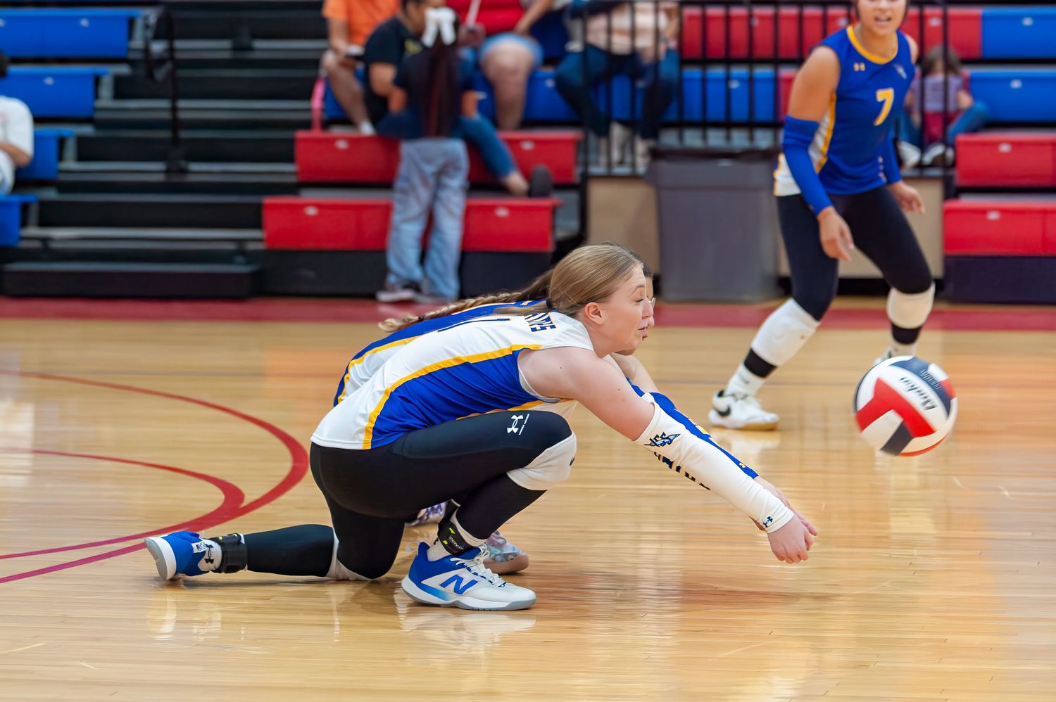 Female volleyball player diving to dig a ball during an indoor match, wearing blue and gold uniform.
