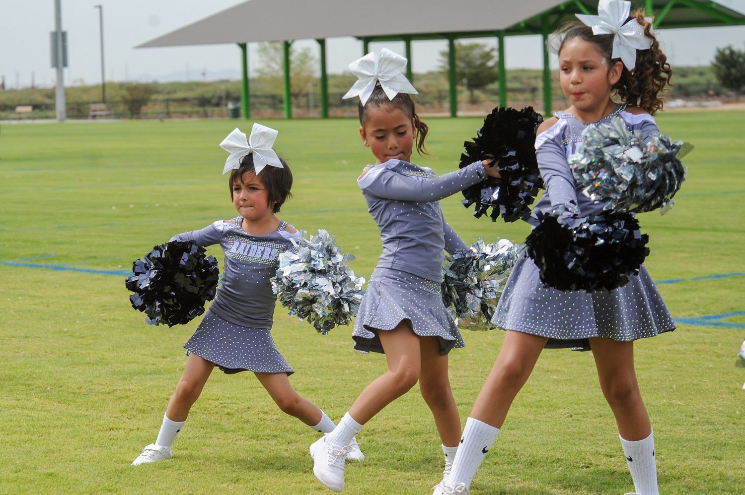 Three young girl cheerleaders in gray uniforms perform routine with black and silver pom-poms on green field.