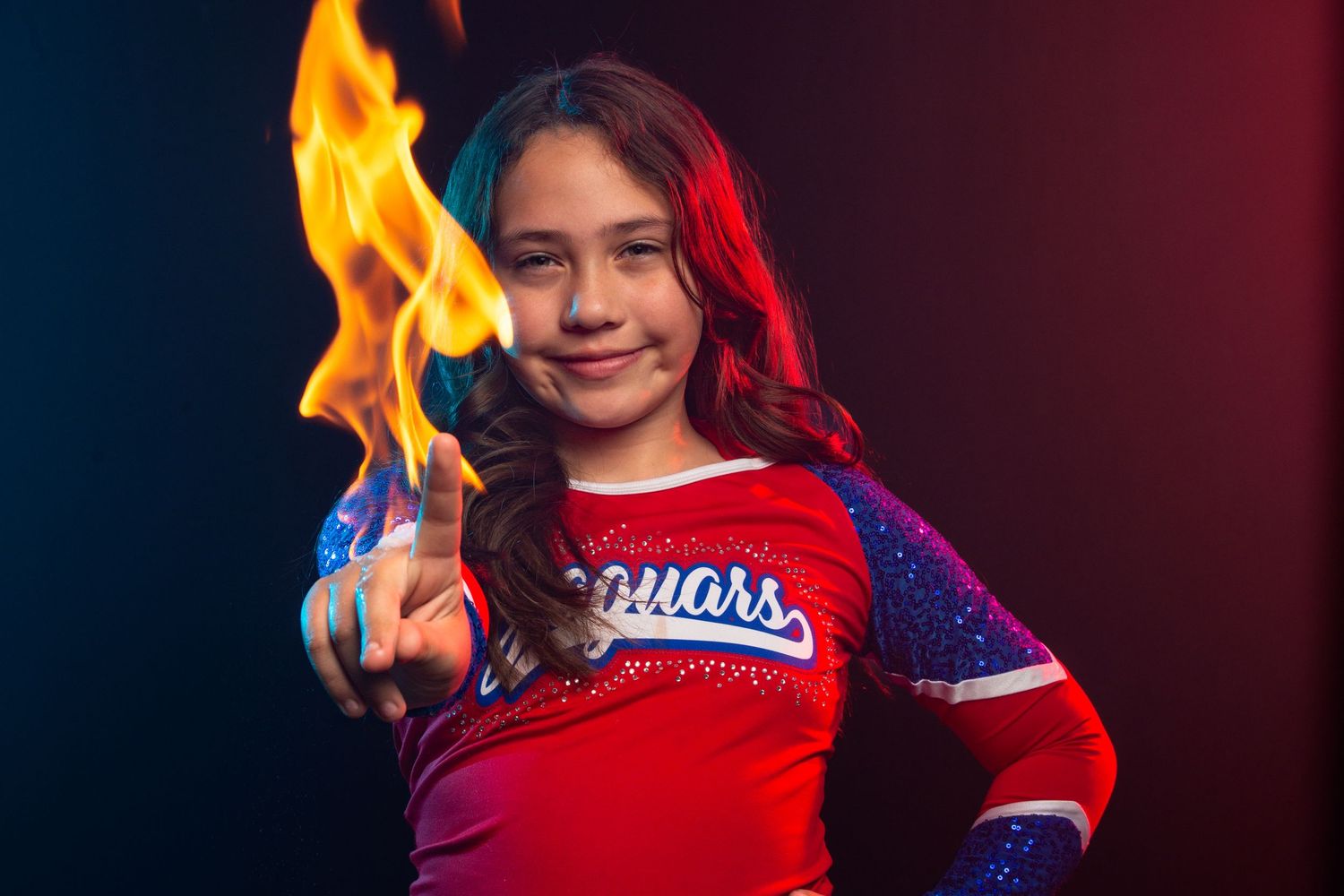 Young girl in Jaguars cheerleader uniform poses confidently with flame effect in dramatic red and blue studio lighting.