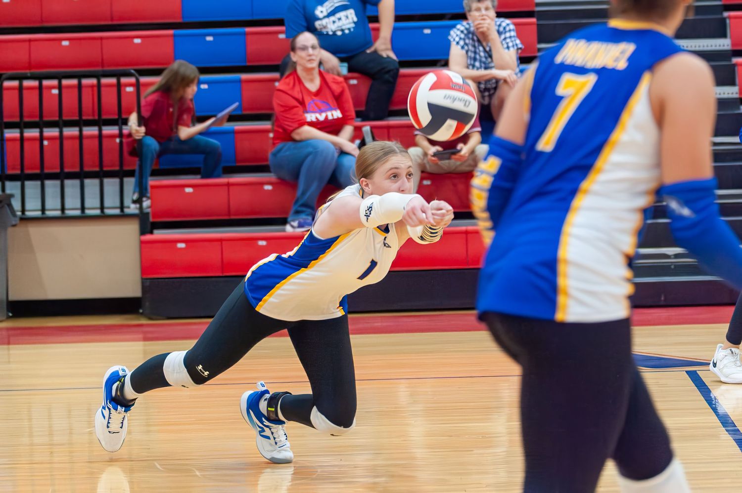 Female volleyball player in blue and gold uniform diving to dig a ball during an indoor match.