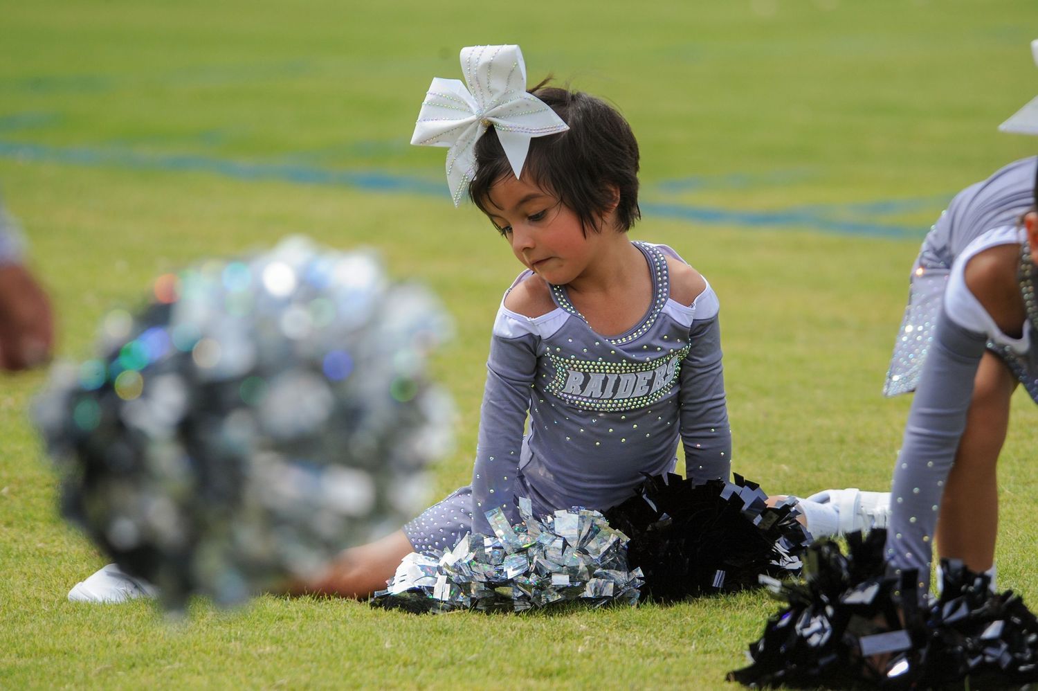 Young cheerleader in silver uniform with white bow sits on grass field holding black and silver pom-poms.