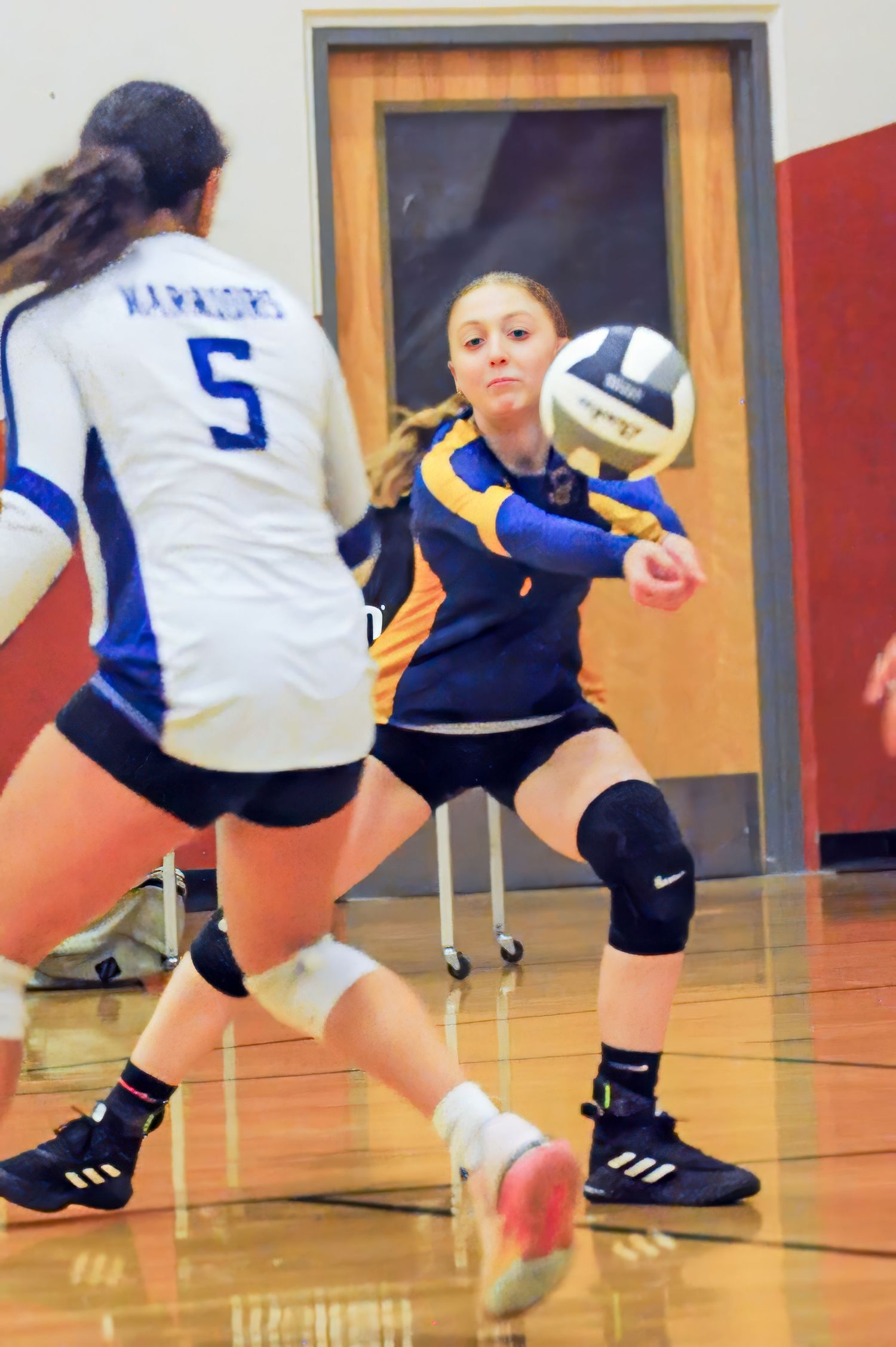 Female volleyball player in blue and gold uniform digs ball during indoor high school match.