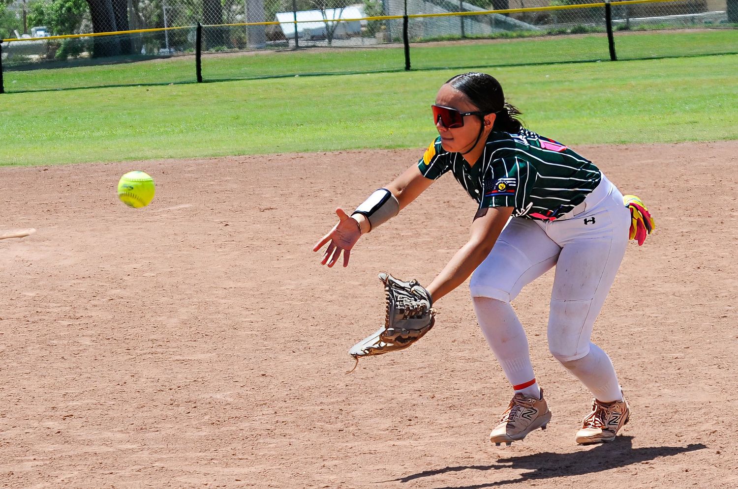 Female softball player in white uniform reaching to field a ground ball on a dirt infield during a game.