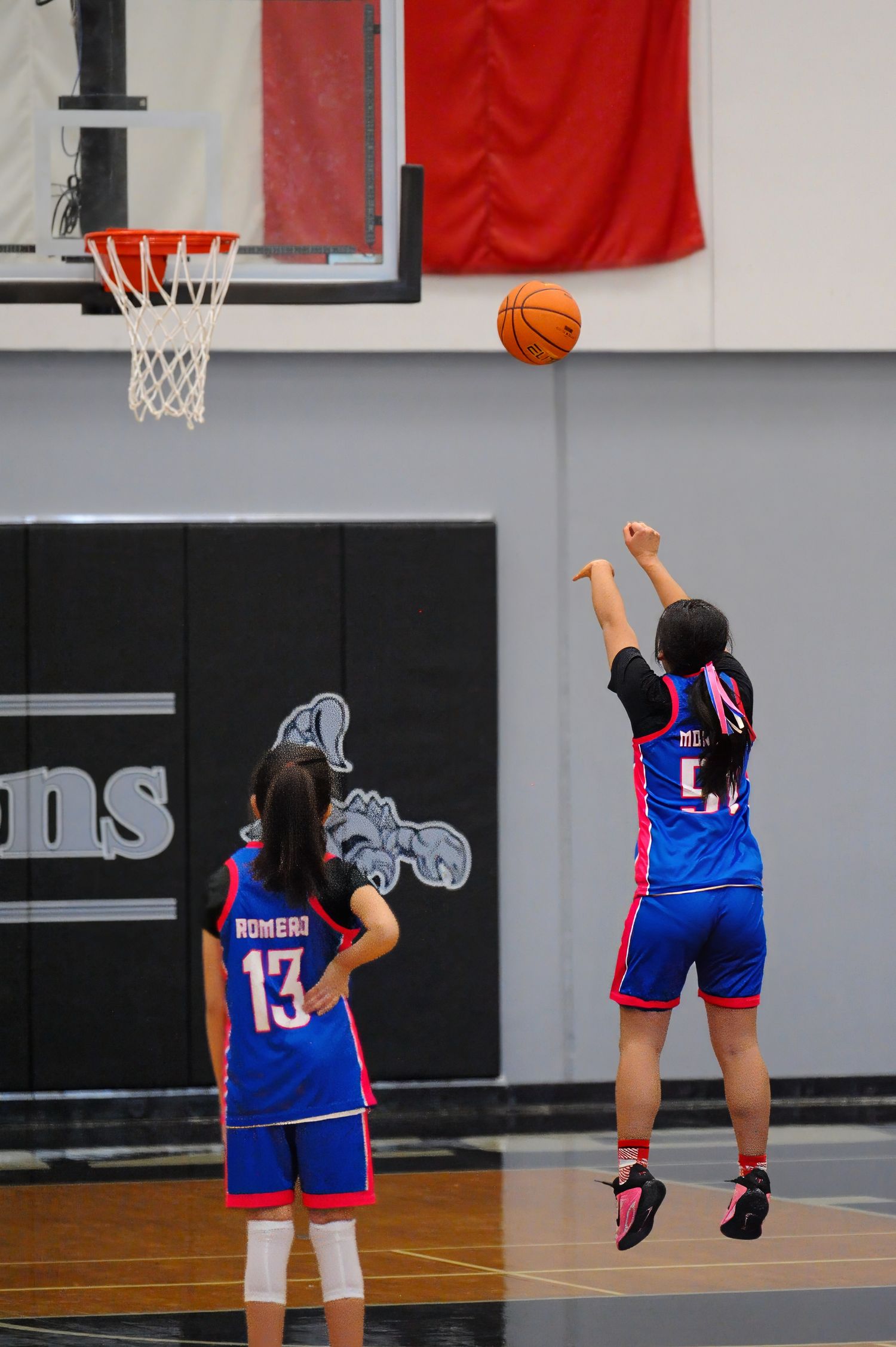 Two girls in blue Romero basketball uniforms, one shooting a jump shot toward the hoop during an indoor youth basketball game.