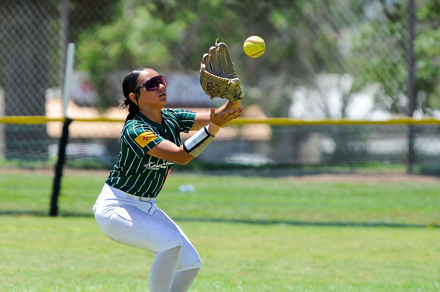 Female softball player in green uniform crouching to catch a yellow ball with her glove in an outdoor field.
