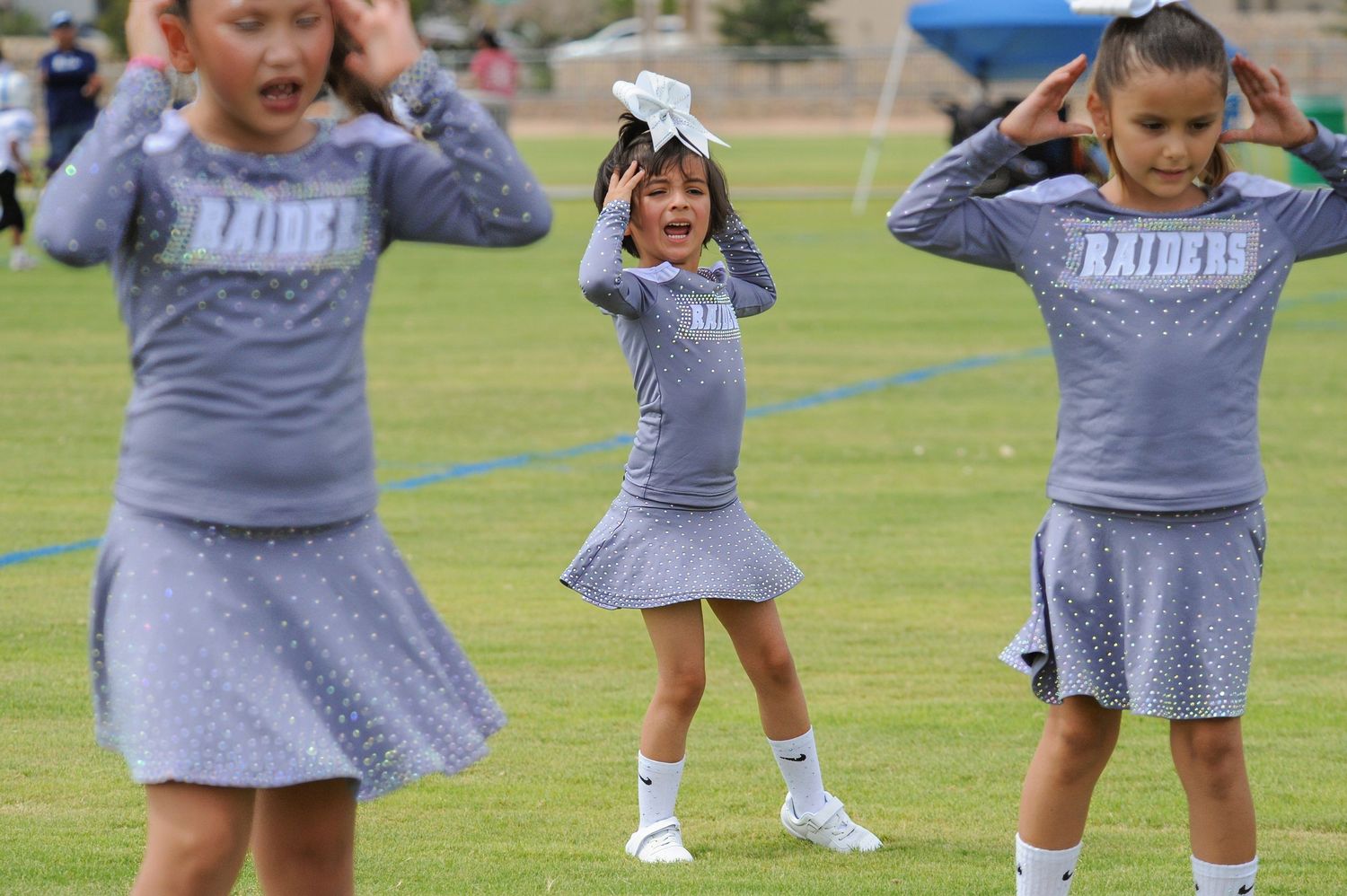 Young girls in Raiders cheerleader uniforms perform a routine on a green field during a youth sports event.