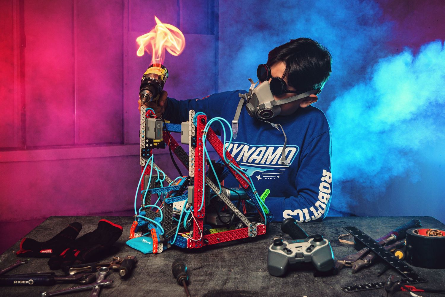 A young engineer wearing a gas mask and Dynamo Robotics shirt works on a flaming mechanical device amid colorful smoke.