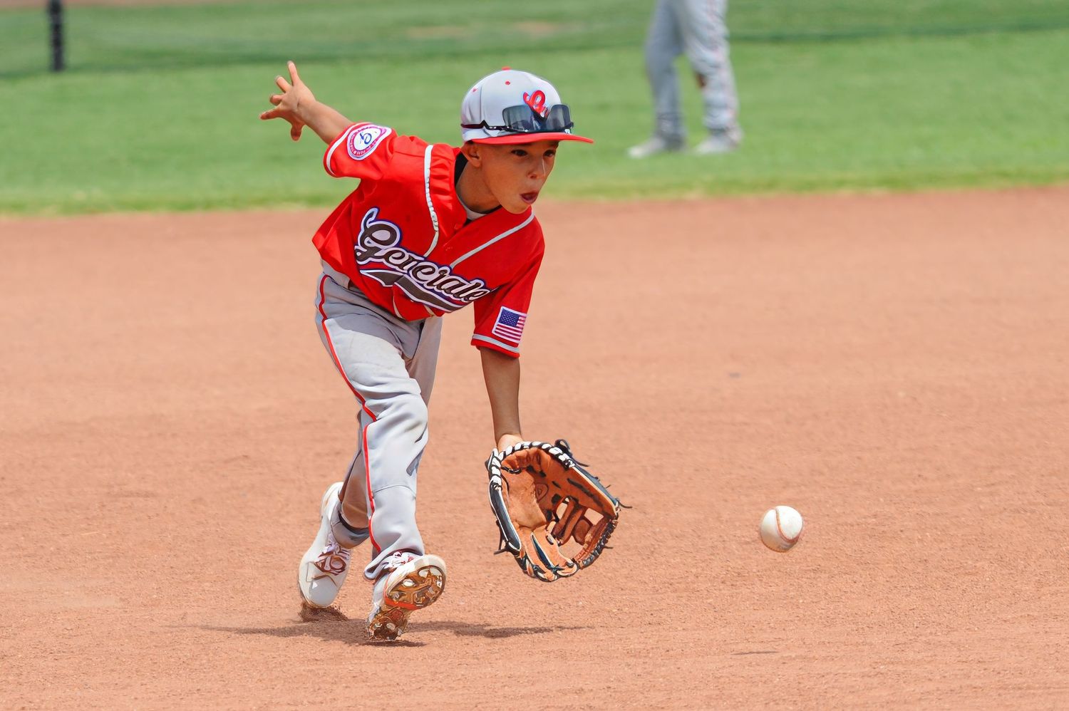 Young baseball player in red uniform fielding ground ball during sunny youth baseball tournament game.