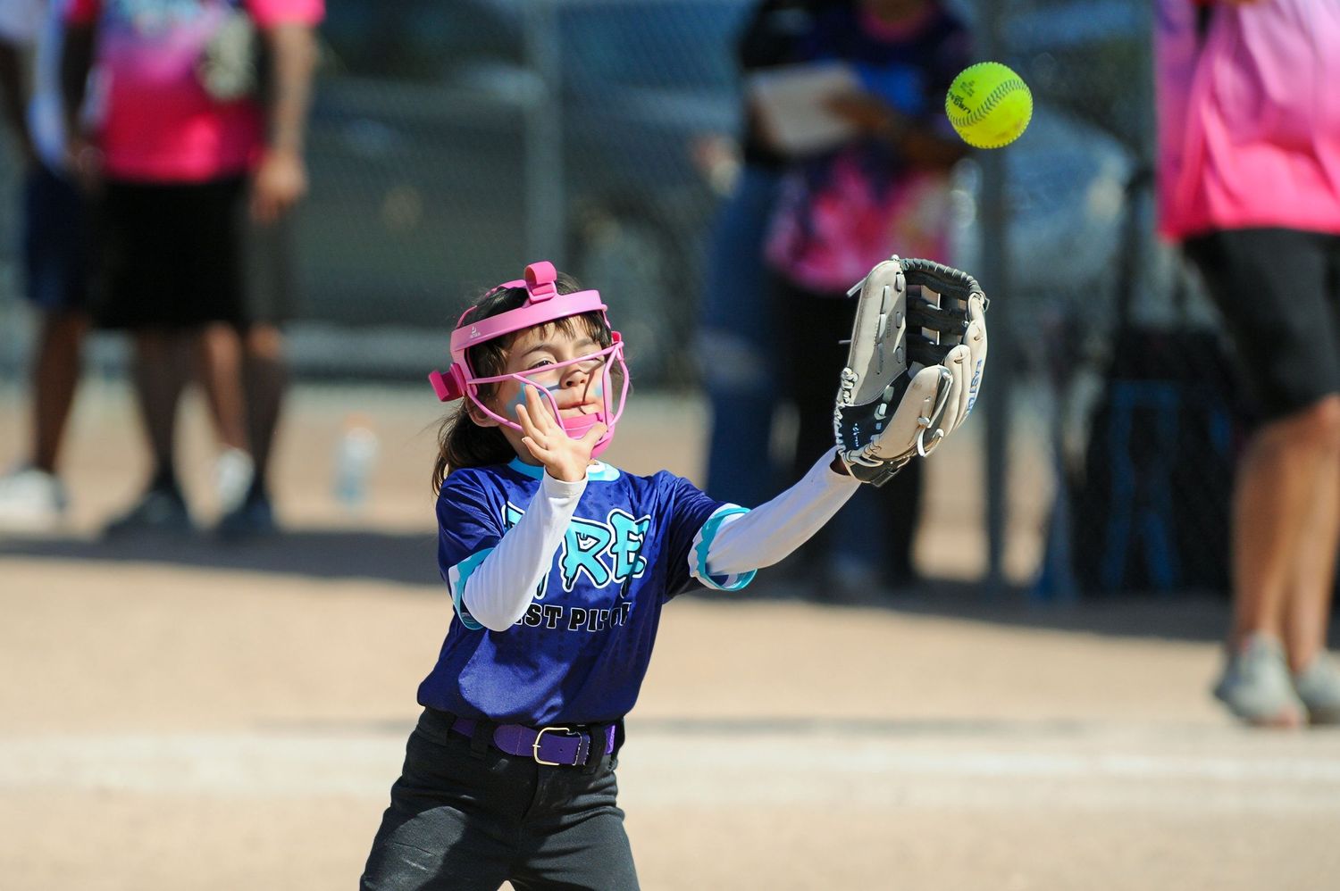 Young softball player in purple uniform and pink helmet catches a yellow softball in her glove.