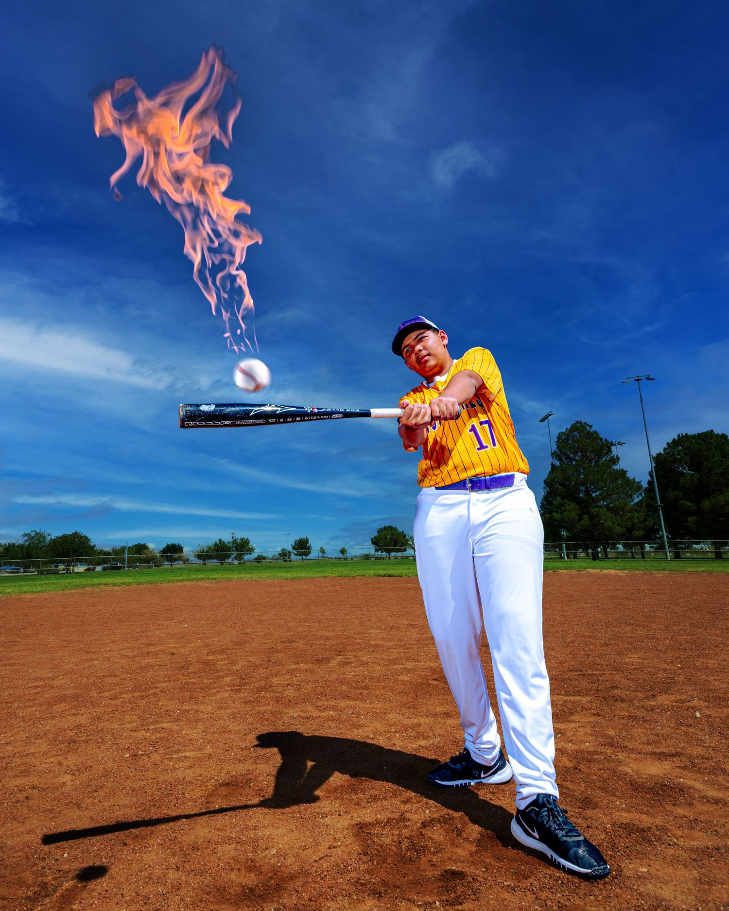 Baseball player in yellow jersey swings bat with flames erupting on contact with ball on sunny outdoor diamond.