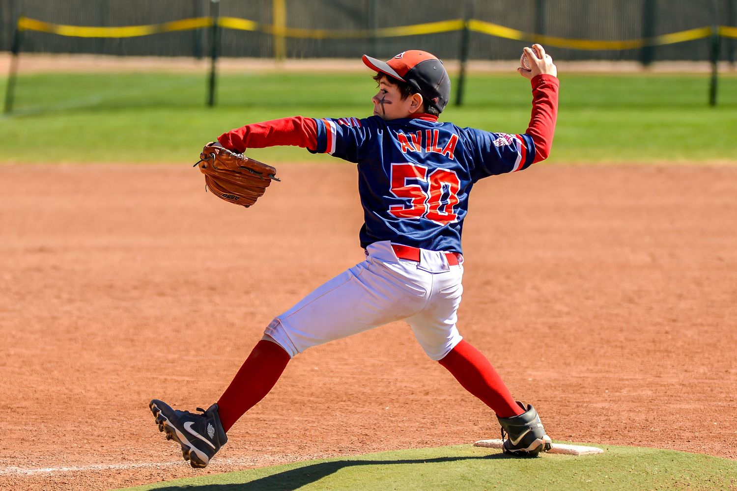 Youth baseball pitcher number 50 in red and blue uniform delivers a pitch on a sunny day.