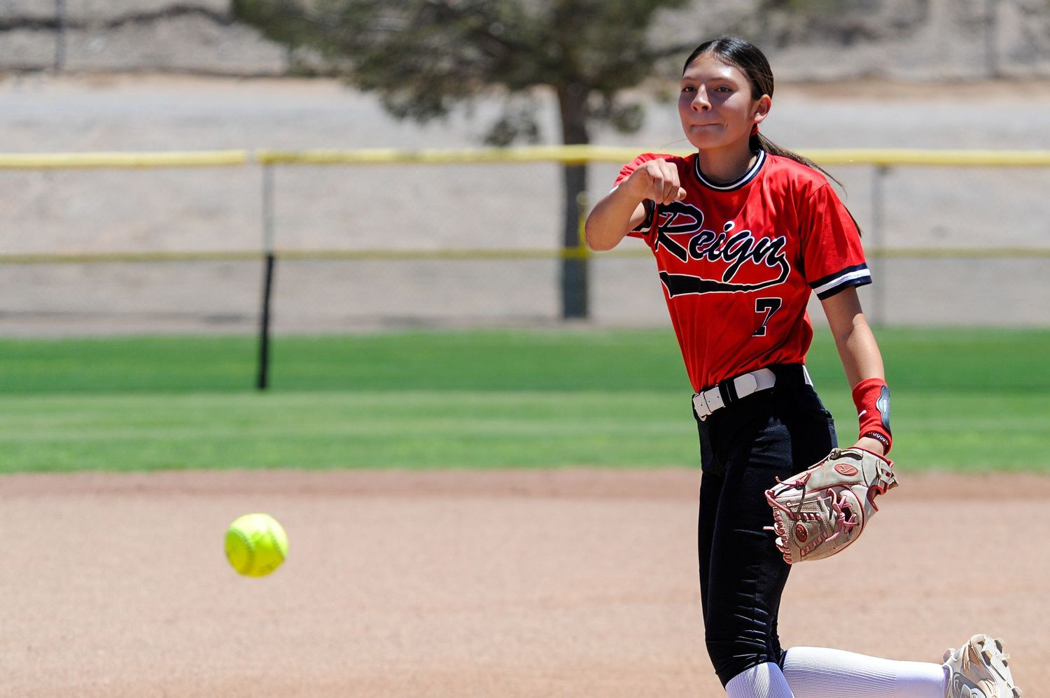 Female softball player in red uniform throwing ball during outdoor game on sunny day.