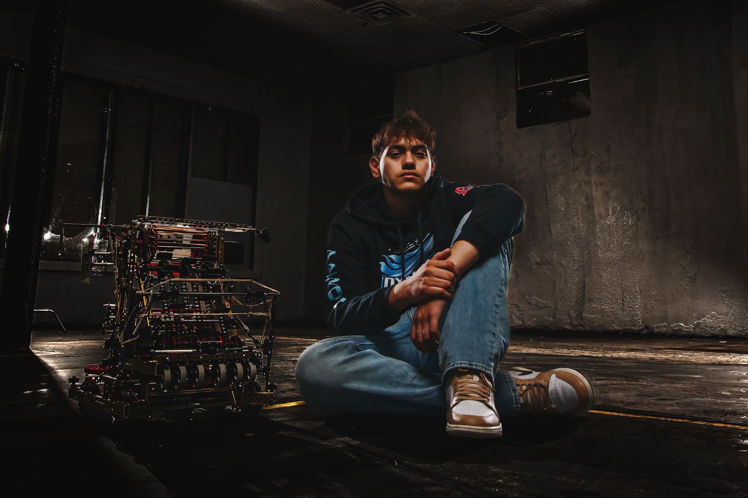 Young man sitting cross-legged on floor in dark studio space with drum kit behind him, wearing black hoodie and jeans.