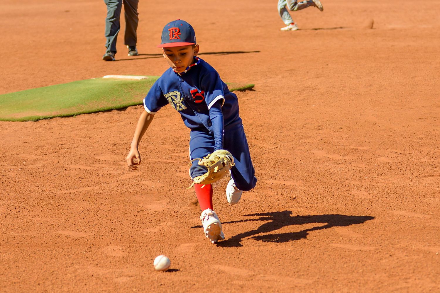 Young baseball player in blue uniform chasing a ground ball across a sun-drenched dirt infield.
