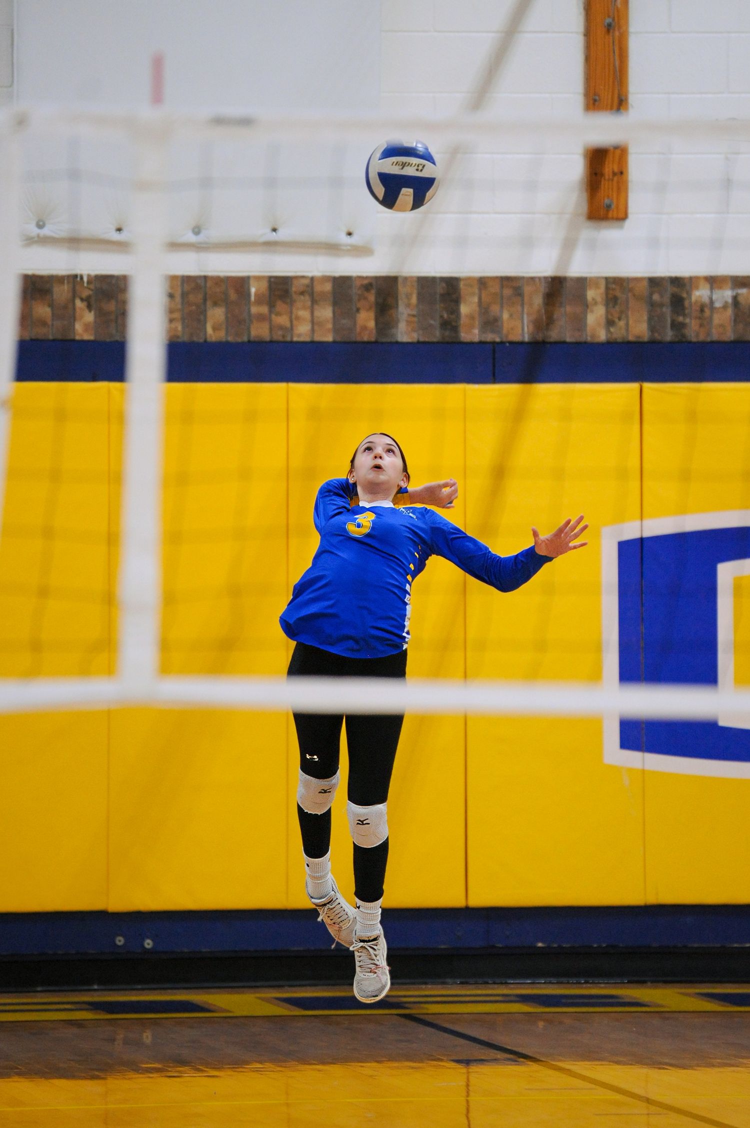 Female volleyball player in blue uniform jumps to spike ball during indoor gym match with yellow and blue walls.
