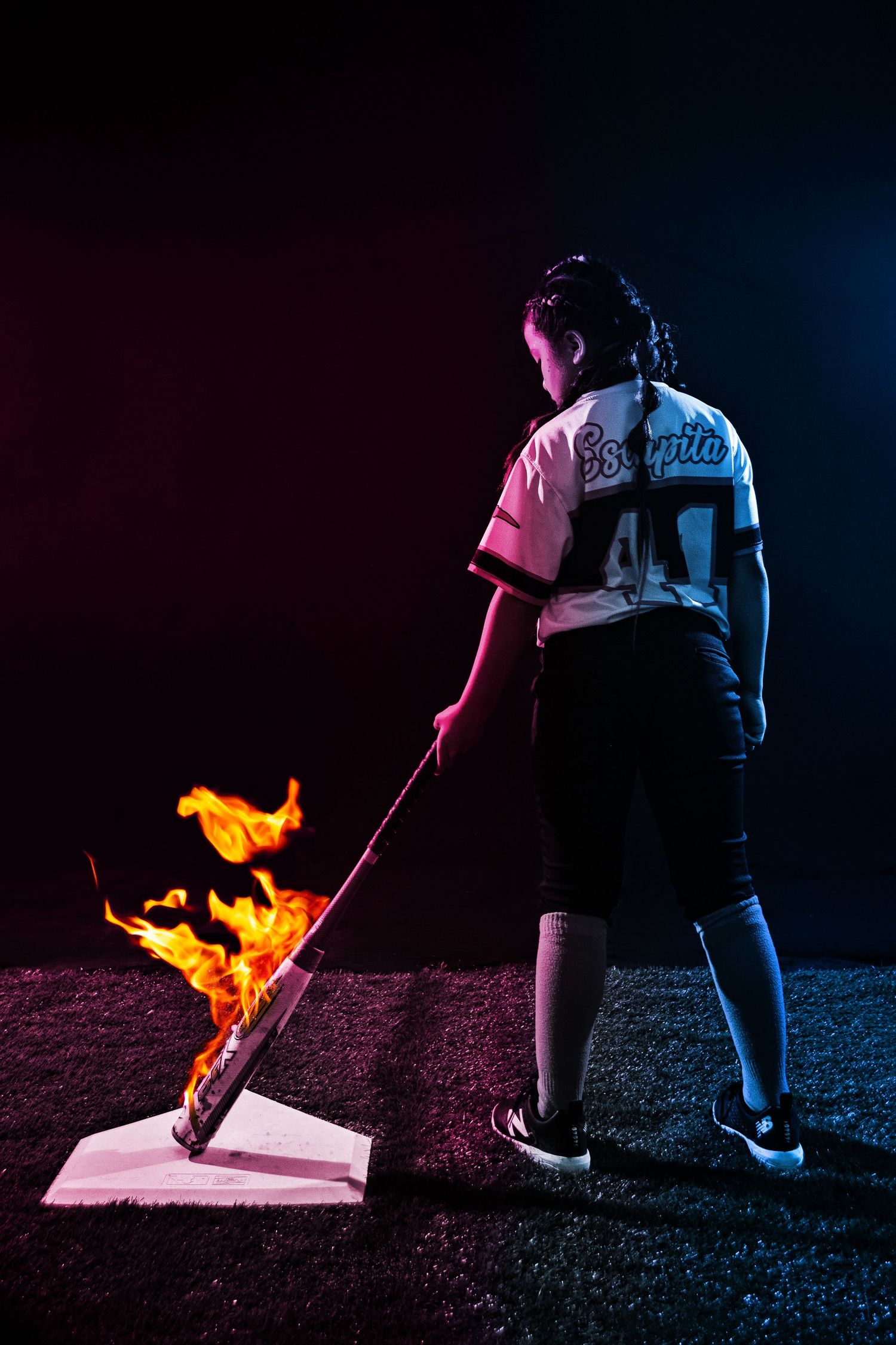Baseball player holding a flaming bat over home plate in dramatic dark studio lighting with red and blue tones.
