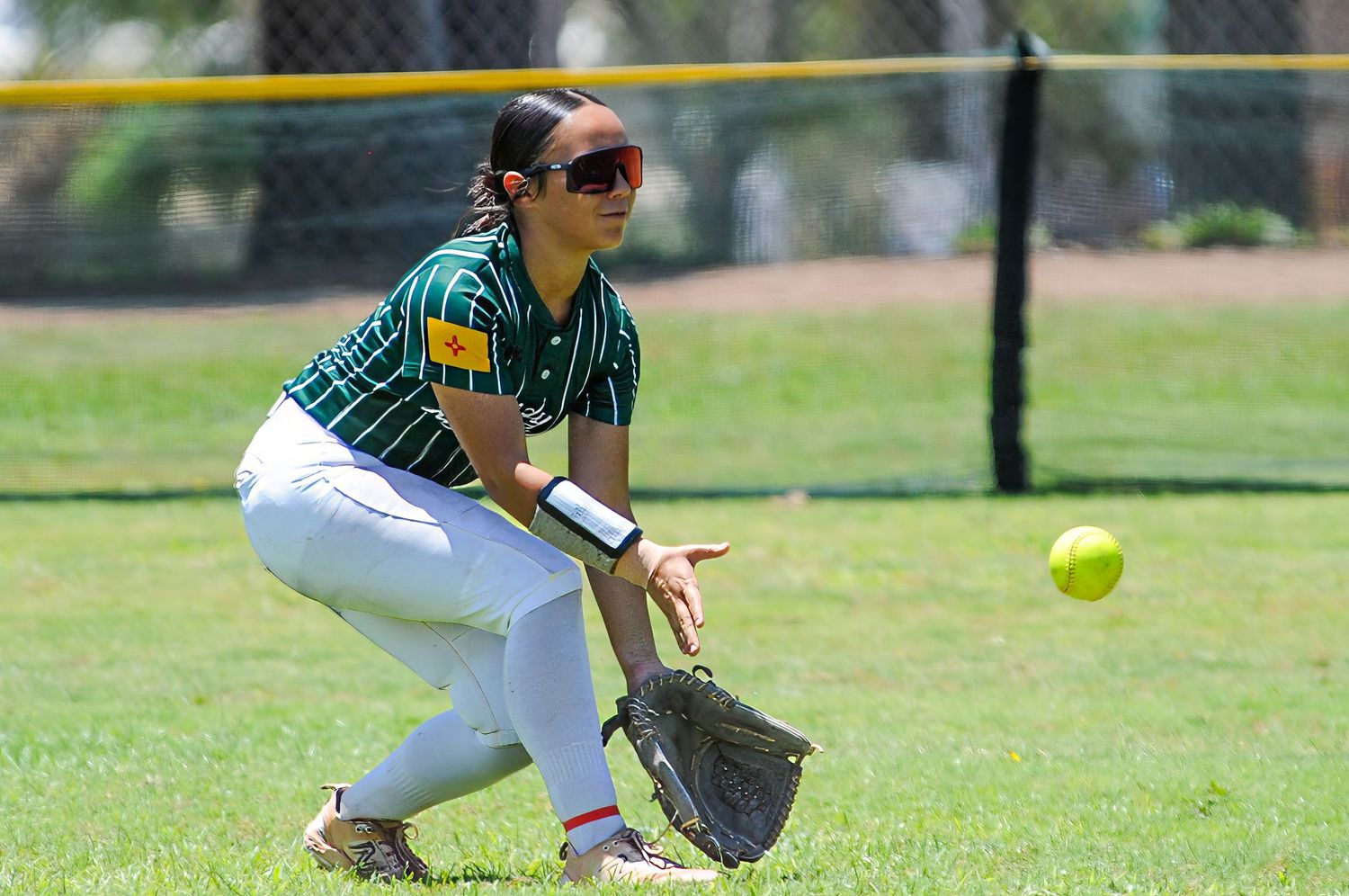 Female softball outfielder in green uniform crouches to field a rolling ball during a sunny game.
