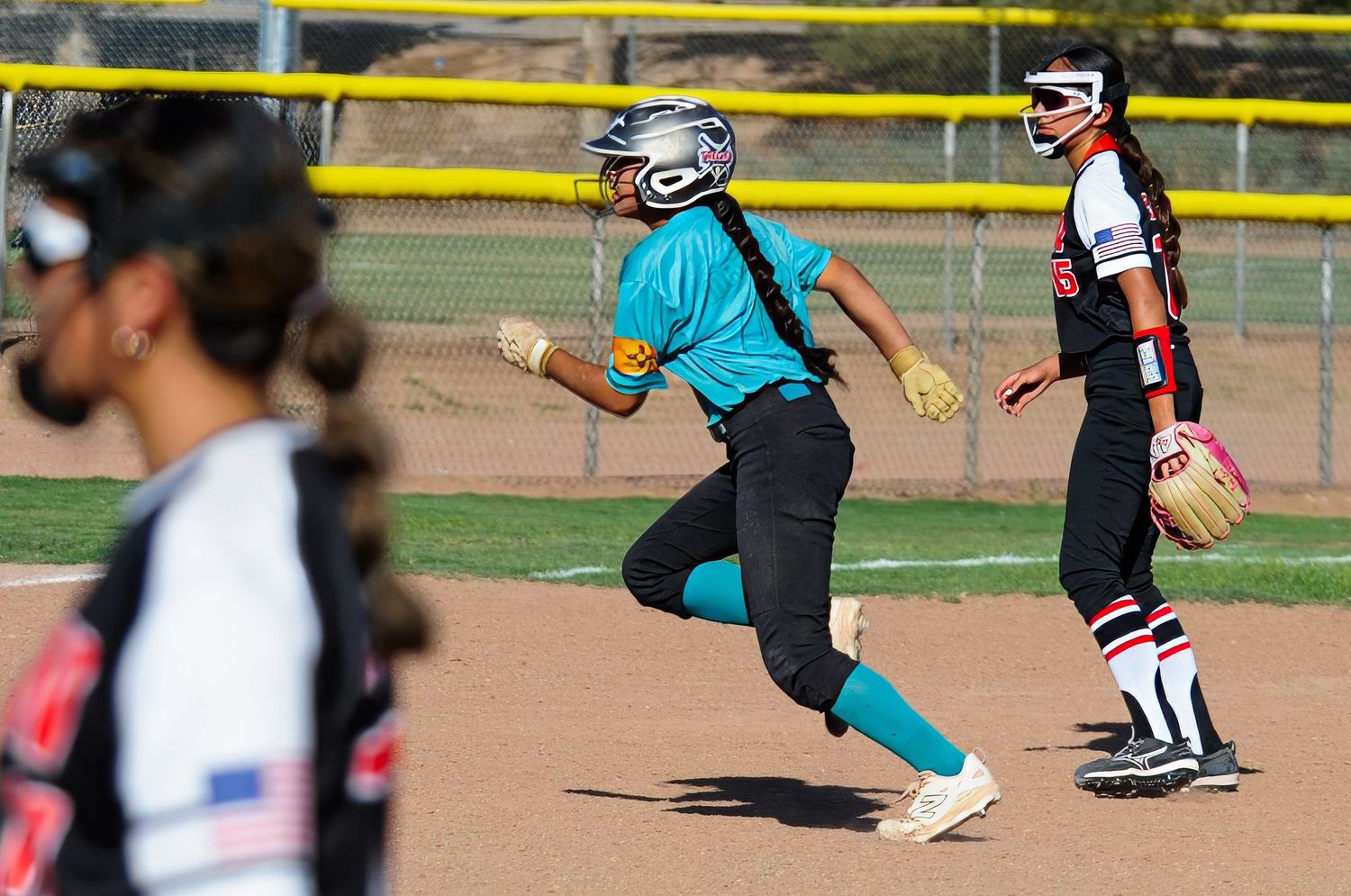 Youth softball player in teal jersey runs bases as opponent in black and white uniform stands nearby.