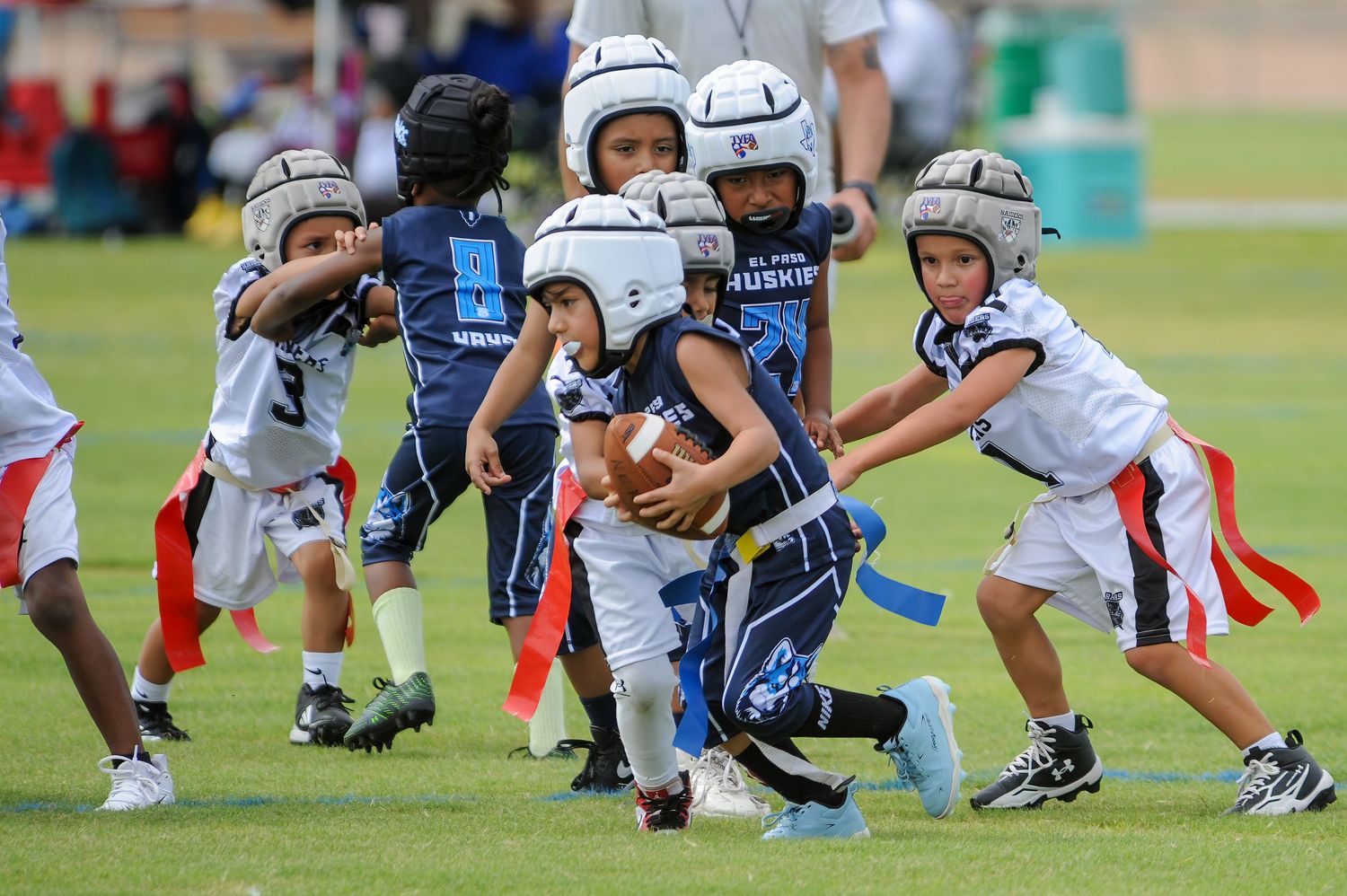 Young children playing flag football in helmets and uniforms during a youth league game on a green field.