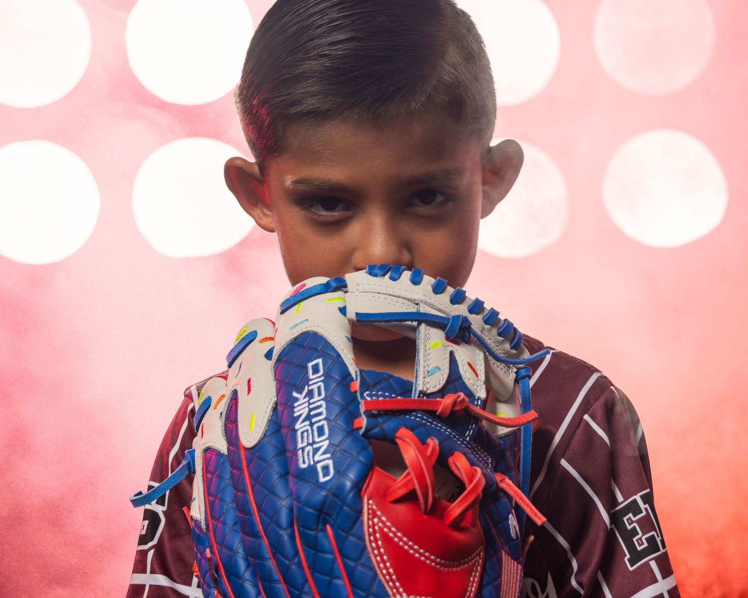 Young baseball player holding a blue and red Diamond baseball glove up to his face against a pink bokeh background.