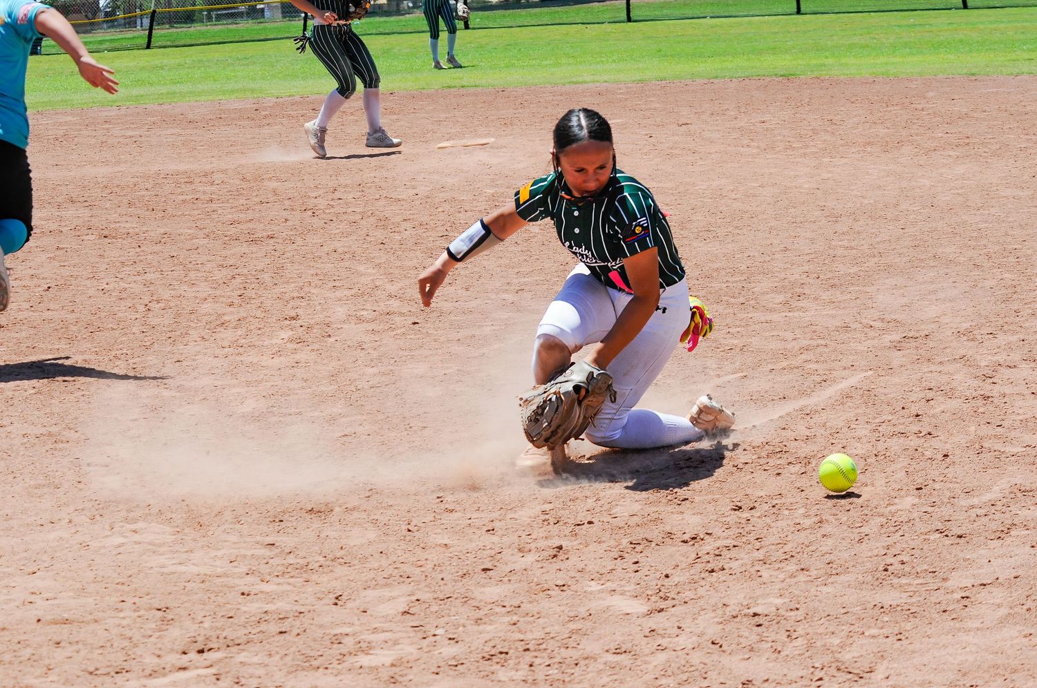 Female softball player sliding on dirt infield attempting to field a yellow ball during an outdoor game.