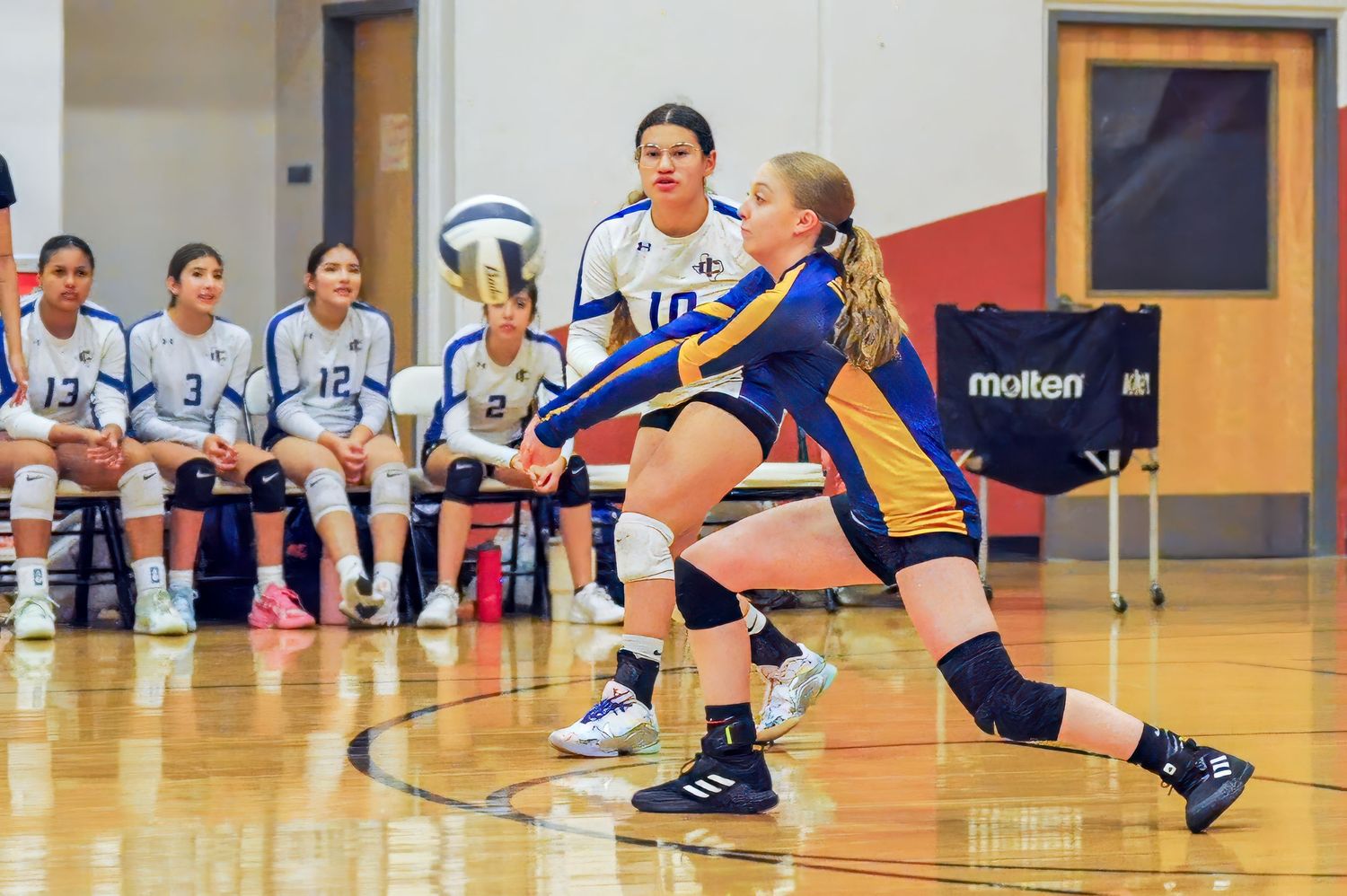 Female volleyball player in gold uniform performs forearm pass during indoor high school volleyball game.