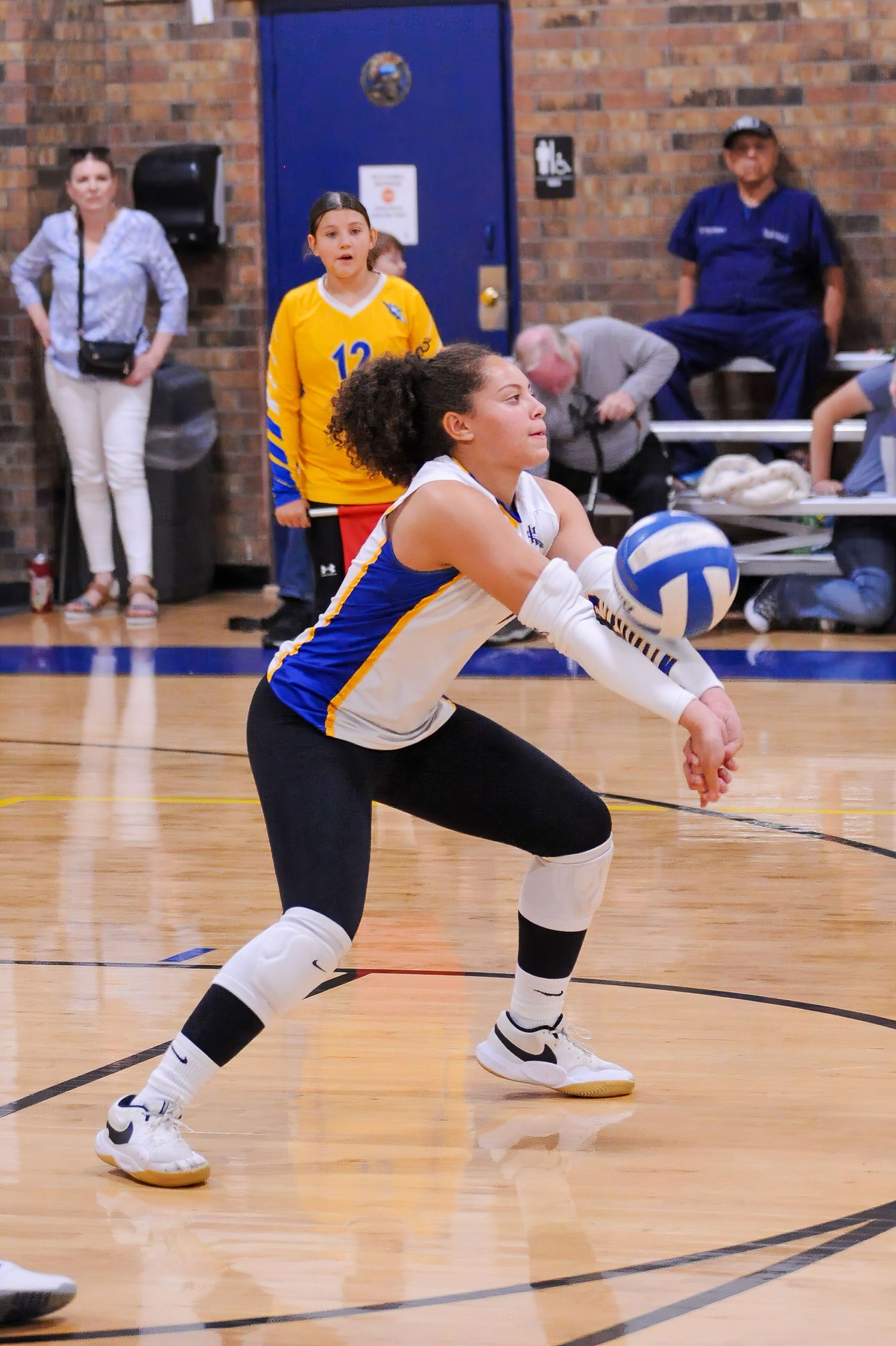 Female volleyball player in blue uniform performs a dig during an indoor volleyball match with spectators watching.
