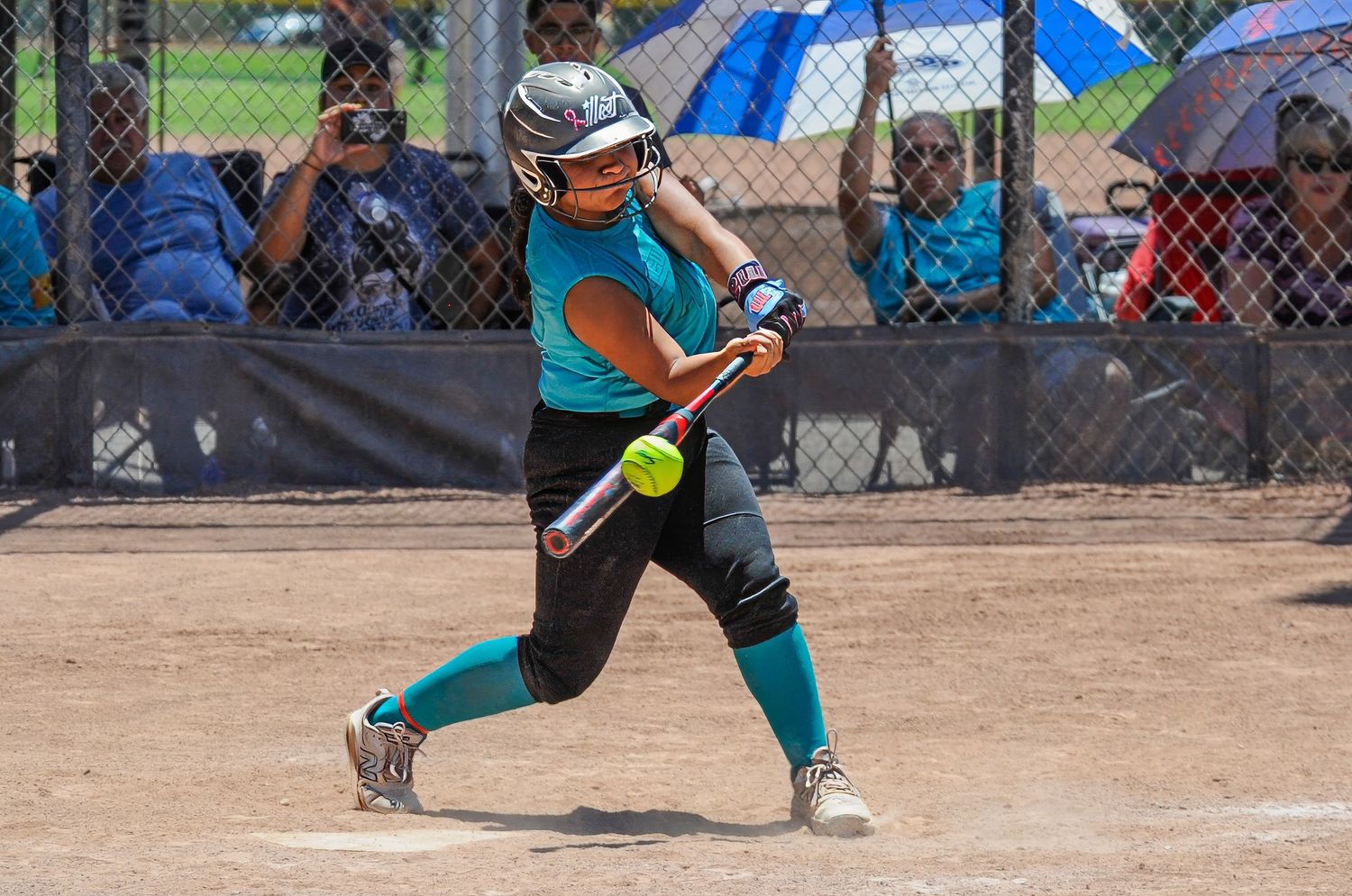 Female softball player in teal uniform swinging bat at yellow ball during sunny outdoor game.