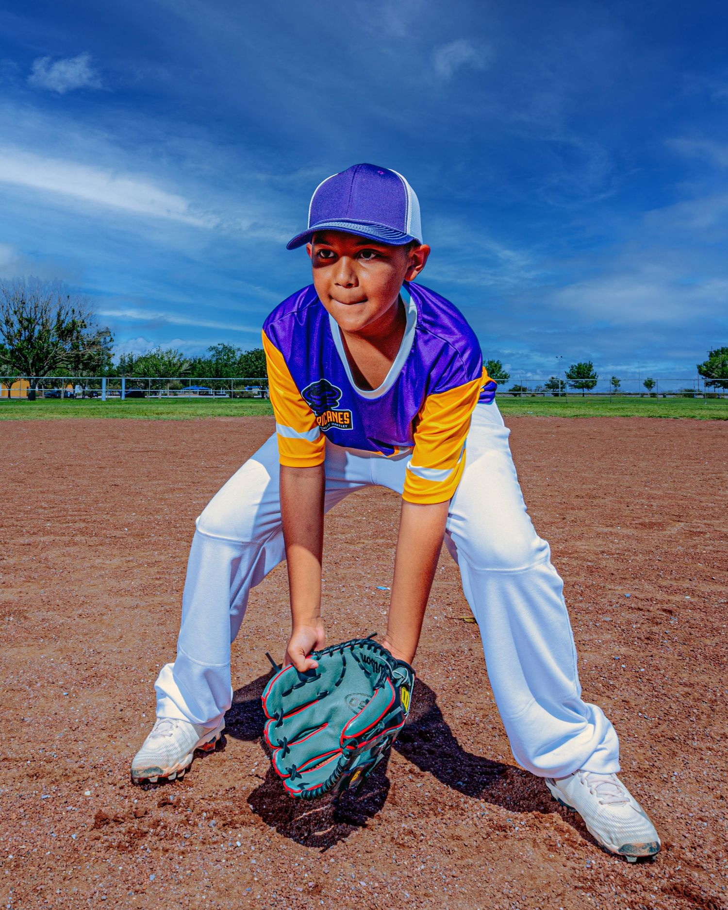 Young baseball player in purple and gold uniform crouches in fielding position with glove on dirt infield.