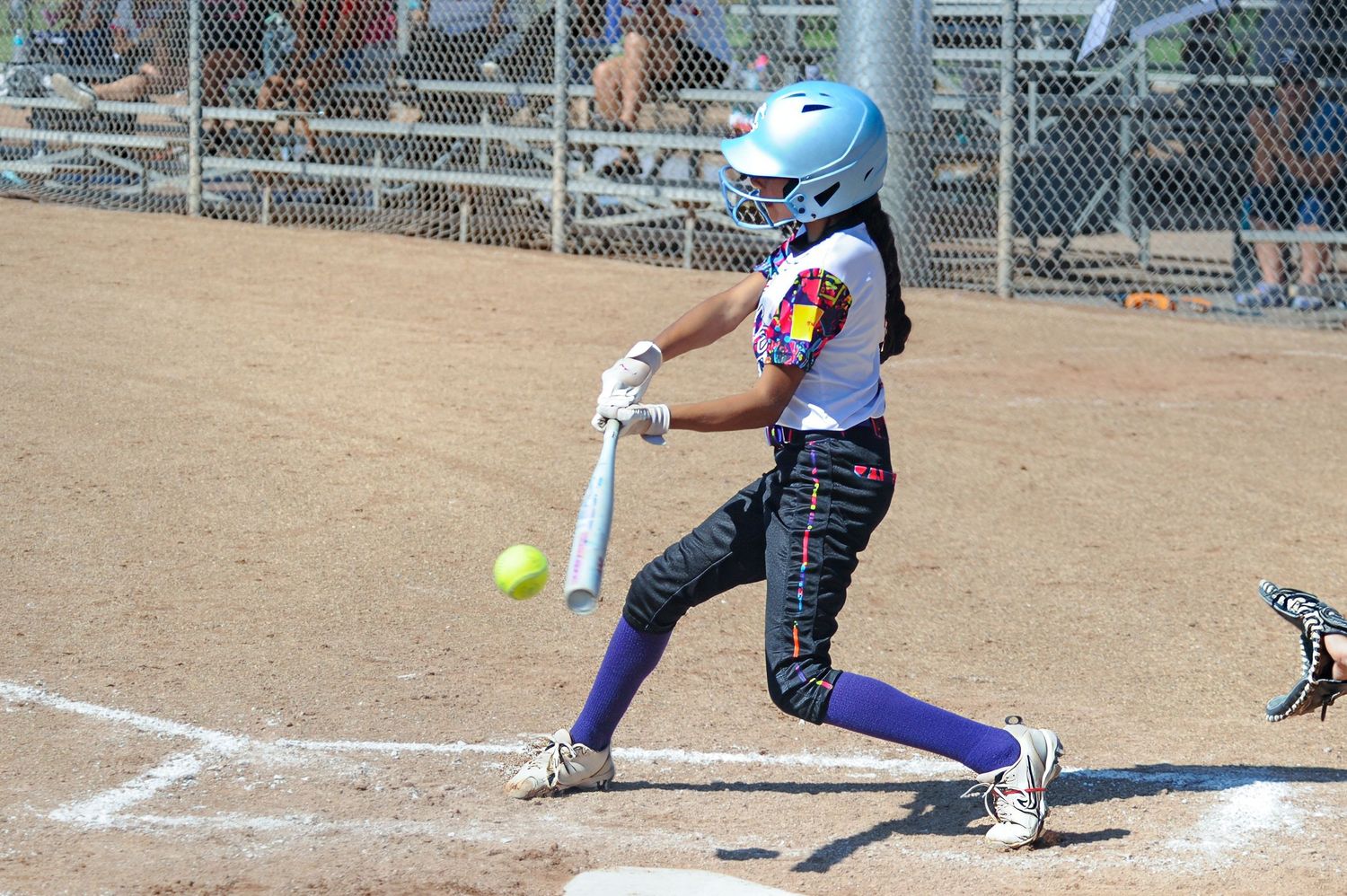 Youth softball player in blue helmet makes contact with a yellow ball during a sunny game.