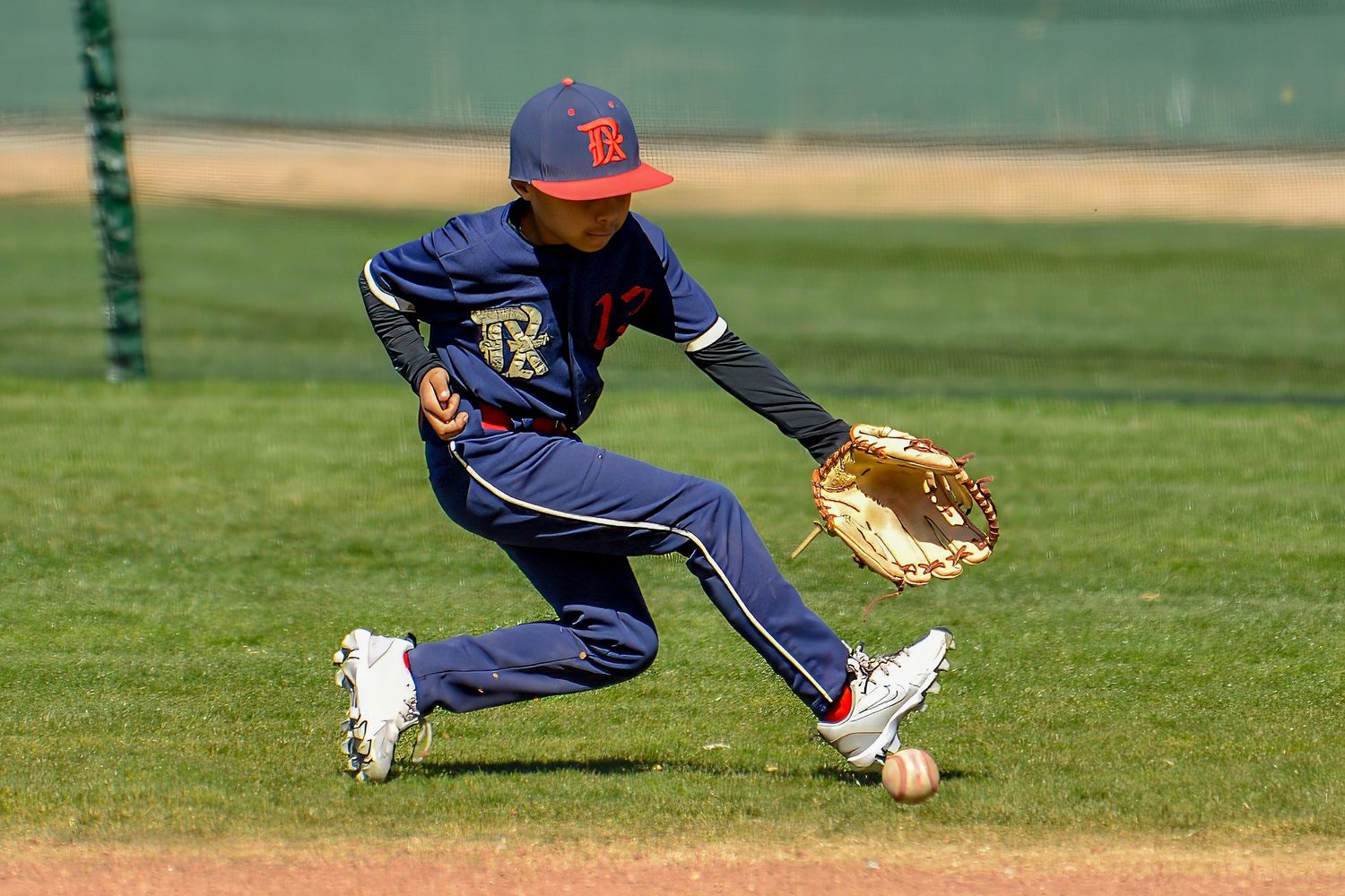 Young baseball player in navy uniform diving low to field a ground ball on a sunny infield.