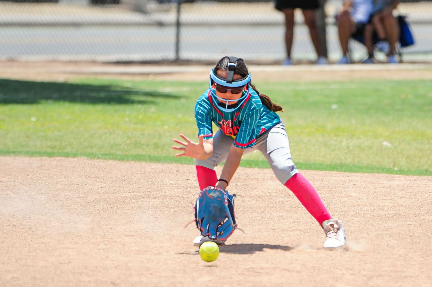Young softball player in teal jersey fields a ground ball during a sunny outdoor youth league game.