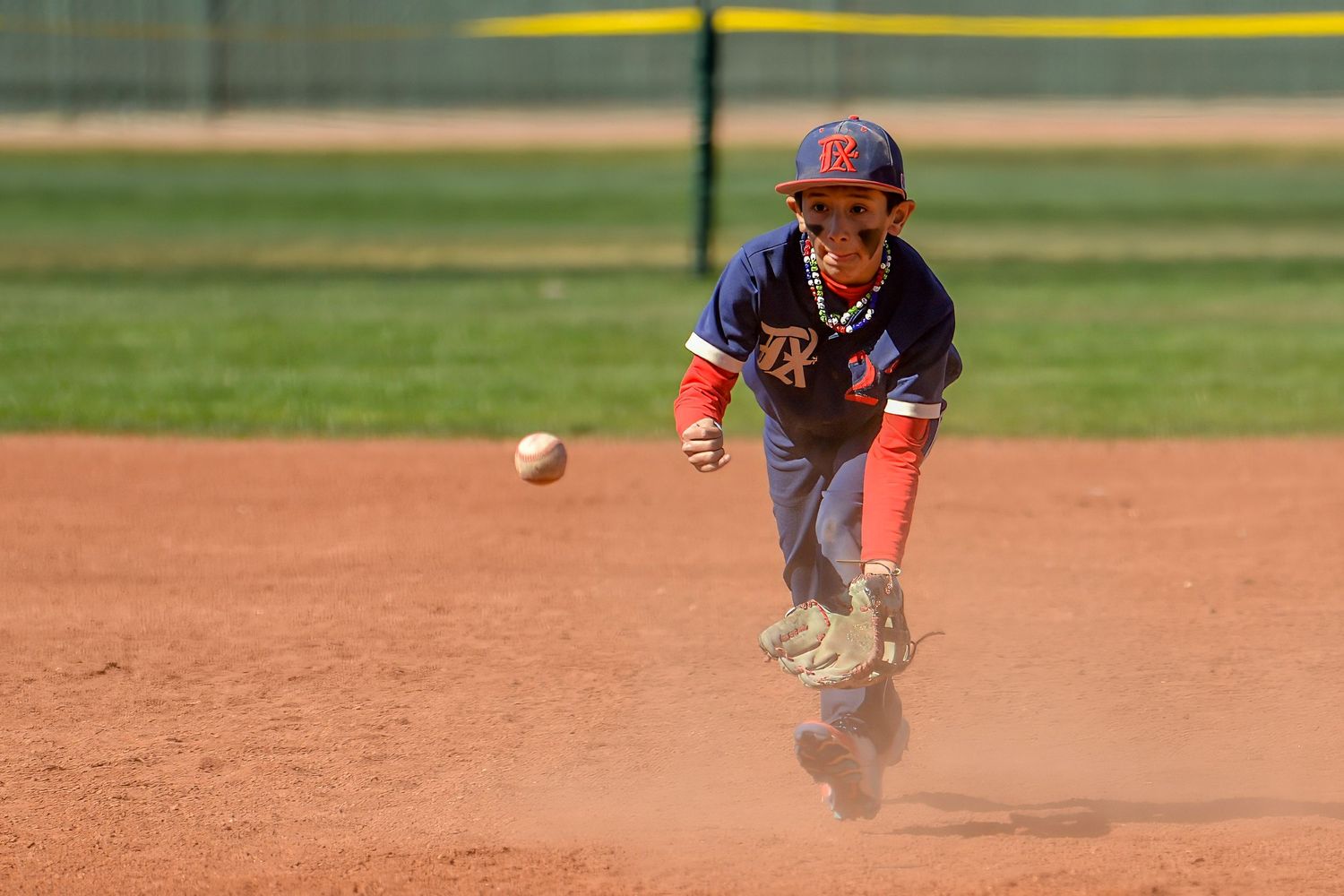 Young baseball player in navy uniform diving to field a rolling ground ball on a dirt infield.