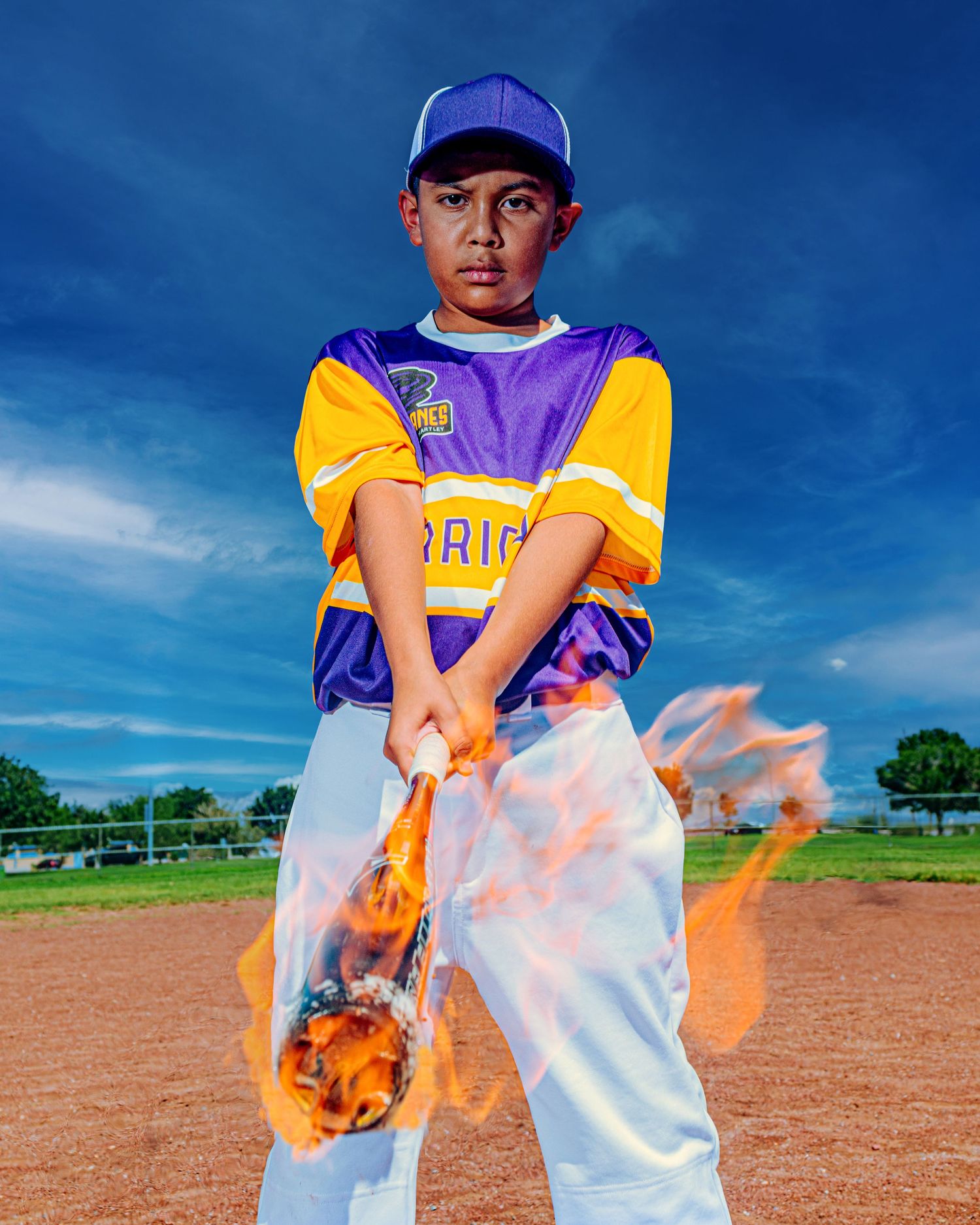 Young baseball player in purple and gold uniform holds a bat with dramatic fire effects on a baseball field.