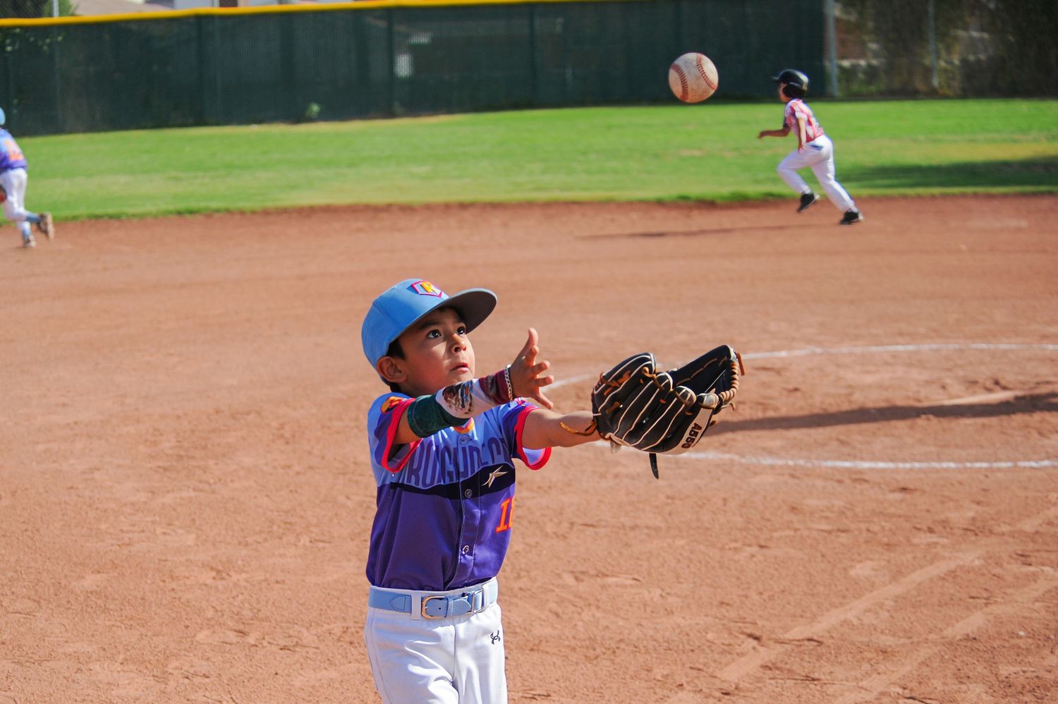 Young baseball player in blue cap and purple jersey reaches up to catch a flying ball with his glove.