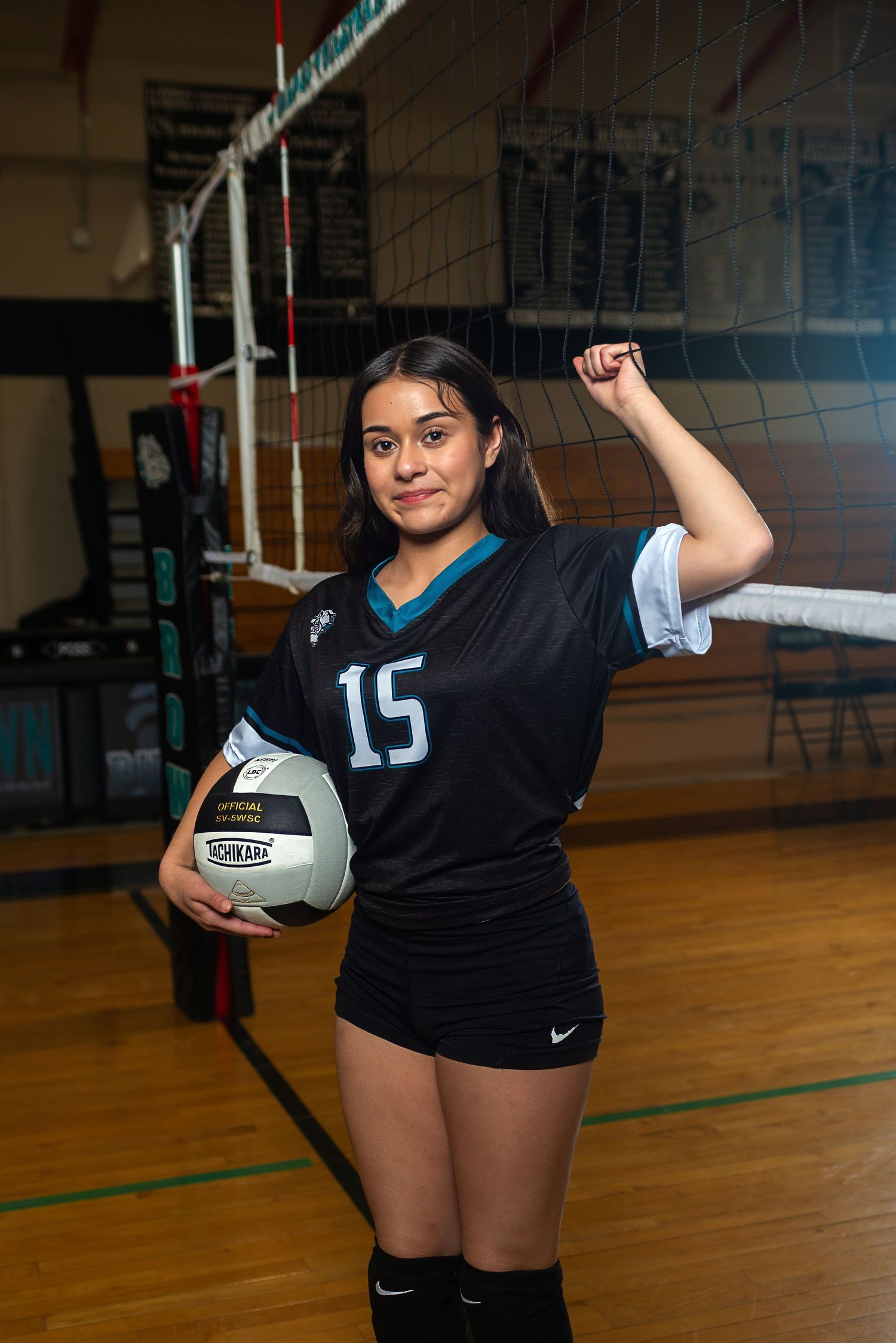 Female volleyball player #15 poses with ball near net in black Nike jersey in gymnasium.