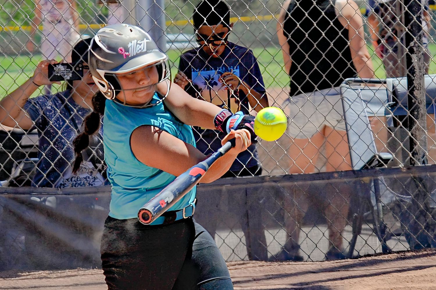 Female softball batter in teal uniform making contact with a yellow pitch during an outdoor game.