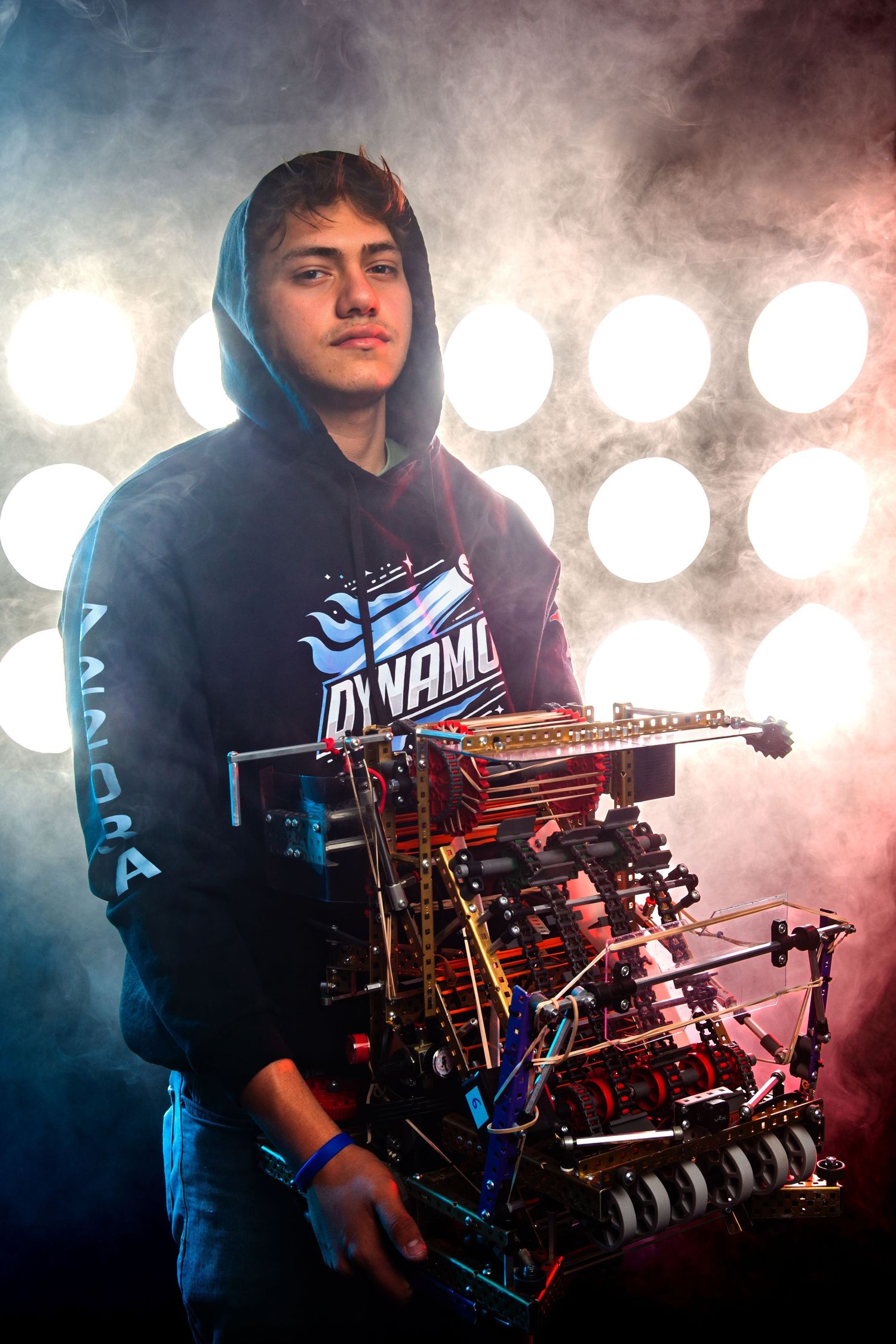 Teen holding a complex robotics competition robot, wearing a Dynamo hoodie, posed dramatically against bright stage lights.