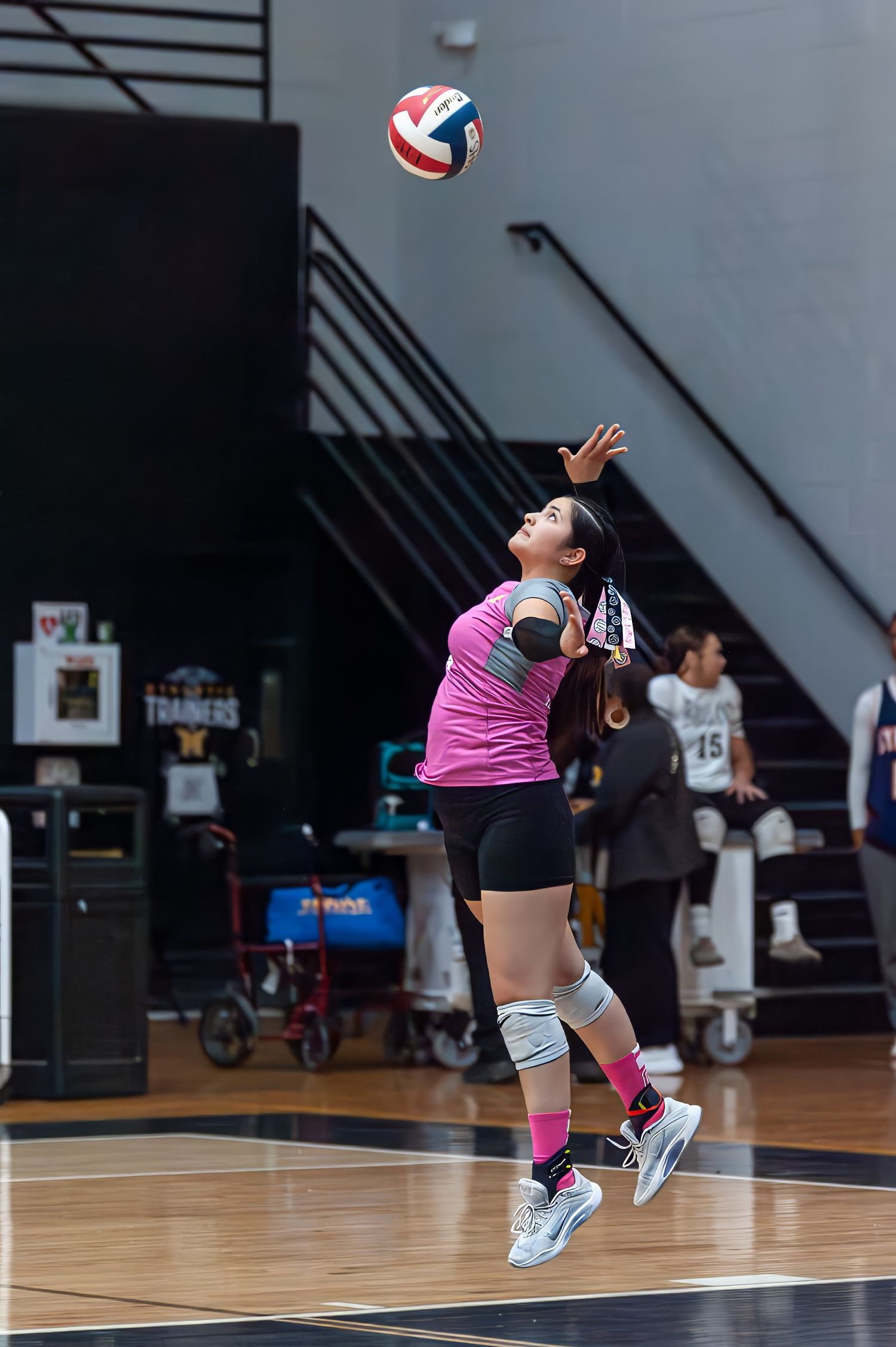 Female volleyball player in pink uniform jumps to serve ball during indoor high school match.