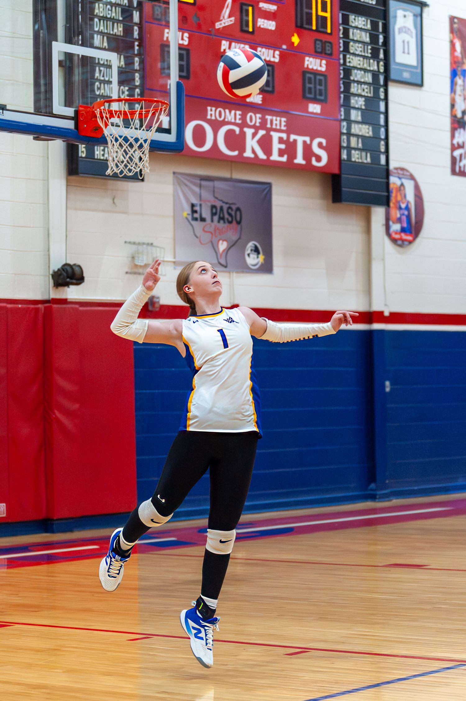 Female volleyball player in white jersey jumping to serve in a school gymnasium with Rockets banner.