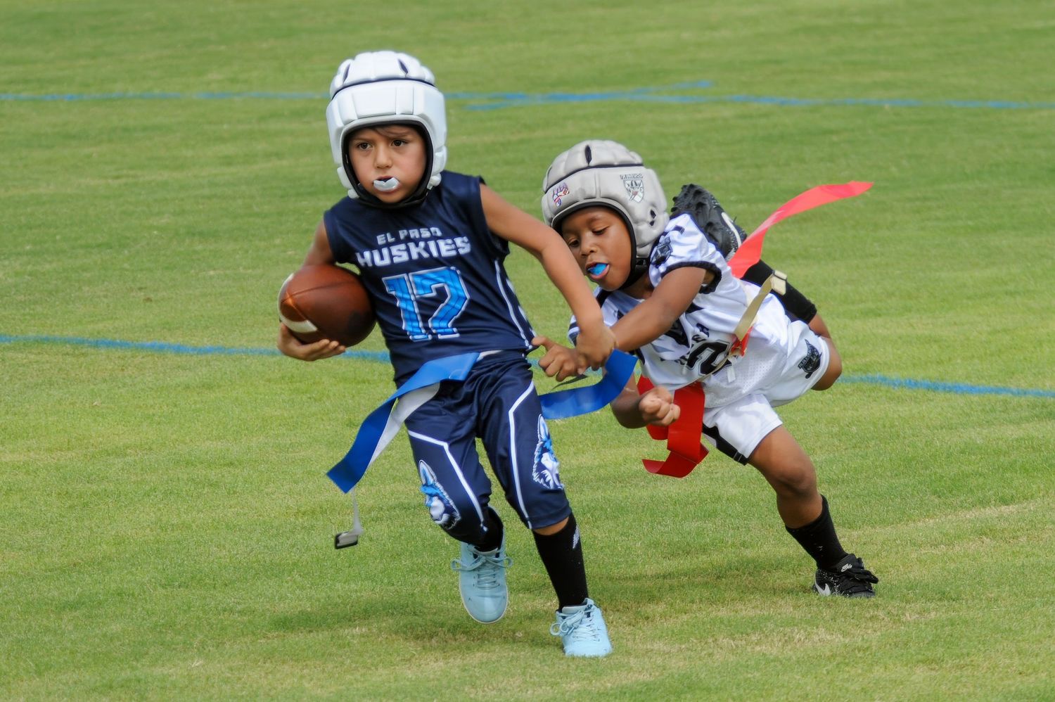 Two young kids playing flag football, one in blue Huskies jersey carrying ball while defender attempts to grab flag.