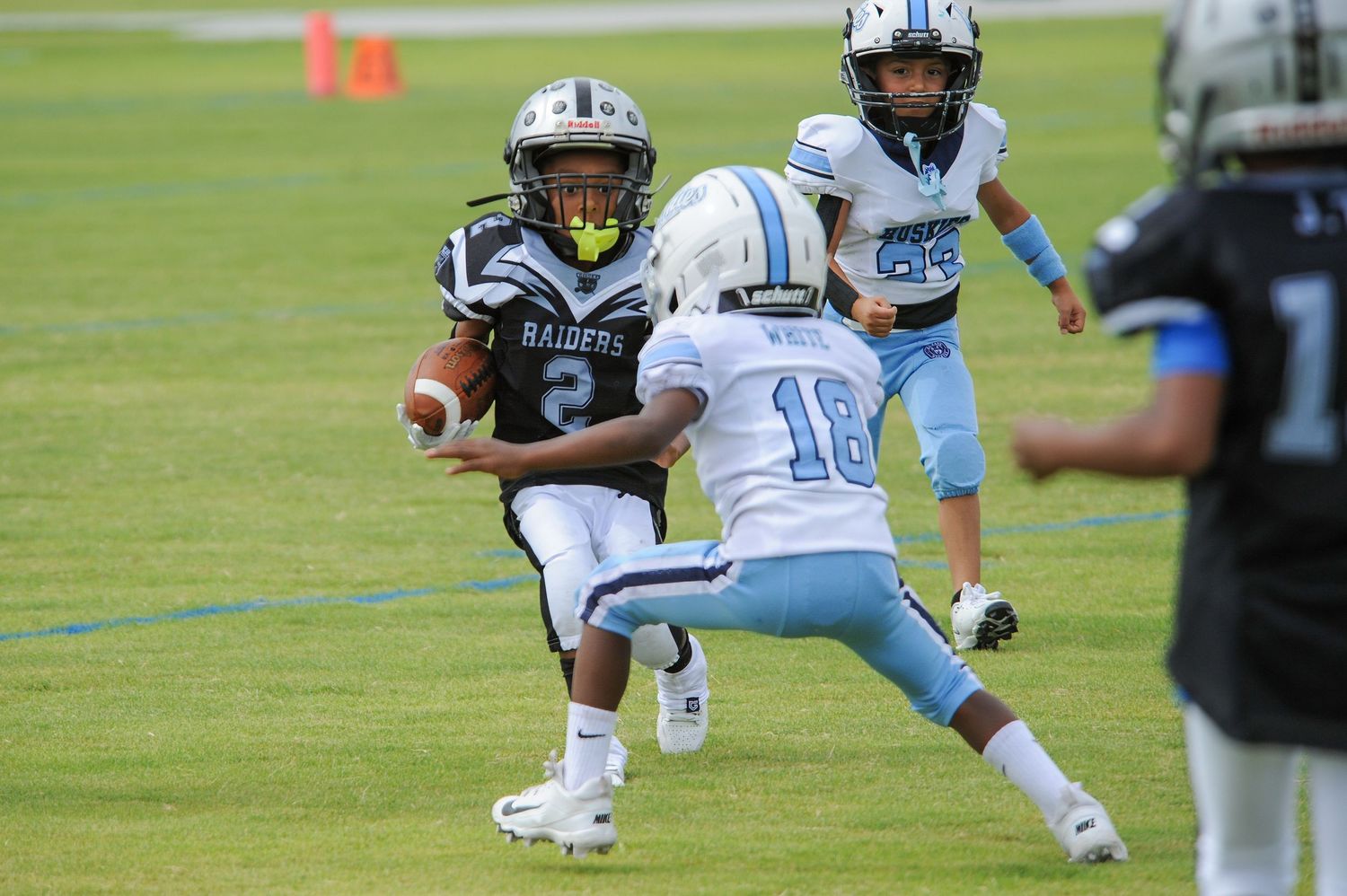 Youth football player in Raiders uniform runs with ball while defender in light blue jersey pursues during youth league game.