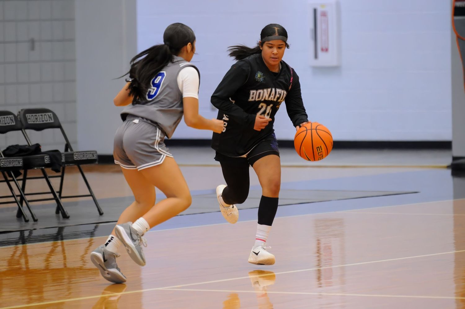 Female basketball player in black Romato jersey dribbles past defender in blue uniform during indoor game.